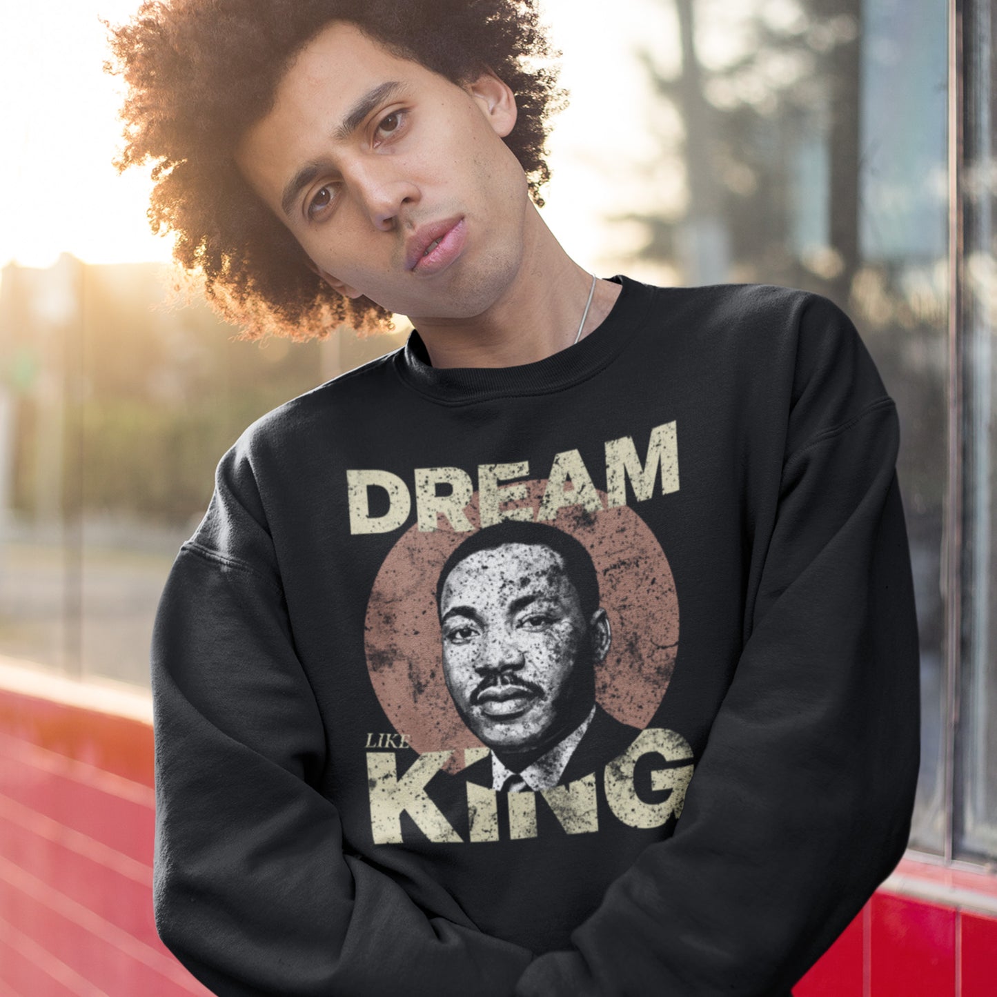 Person wearing a black unisex premium sweatshirt printed with “DREAM LIKE KING” and MLK’s portrait inside a rustic circle. Retro civil-rights artwork in a casual setting.