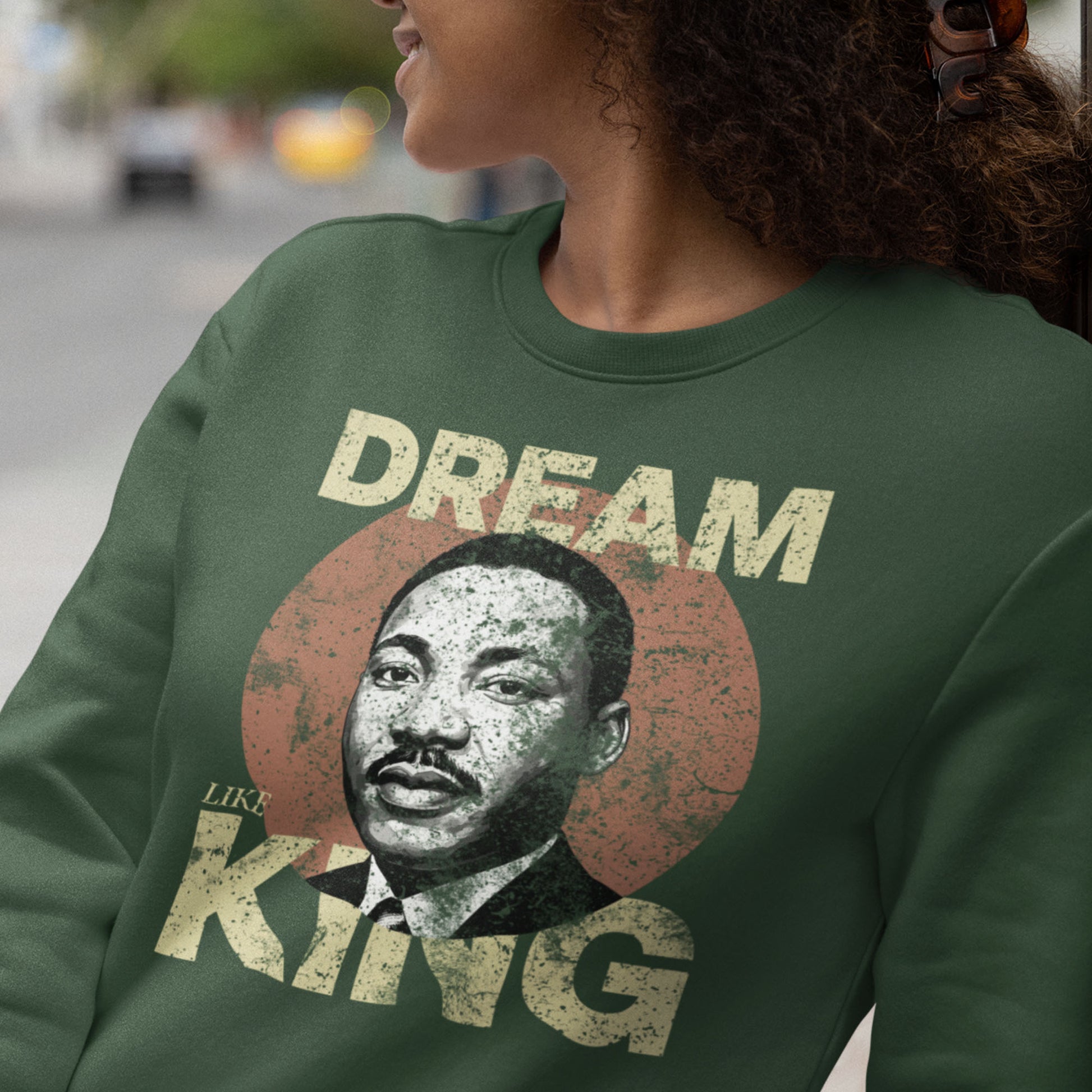 Woman wearing a forest green unisex premium sweatshirt featuring “DREAM LIKE KING” and MLK’s portrait inside a textured circle. Retro civil-rights design with a soft vintage feel.