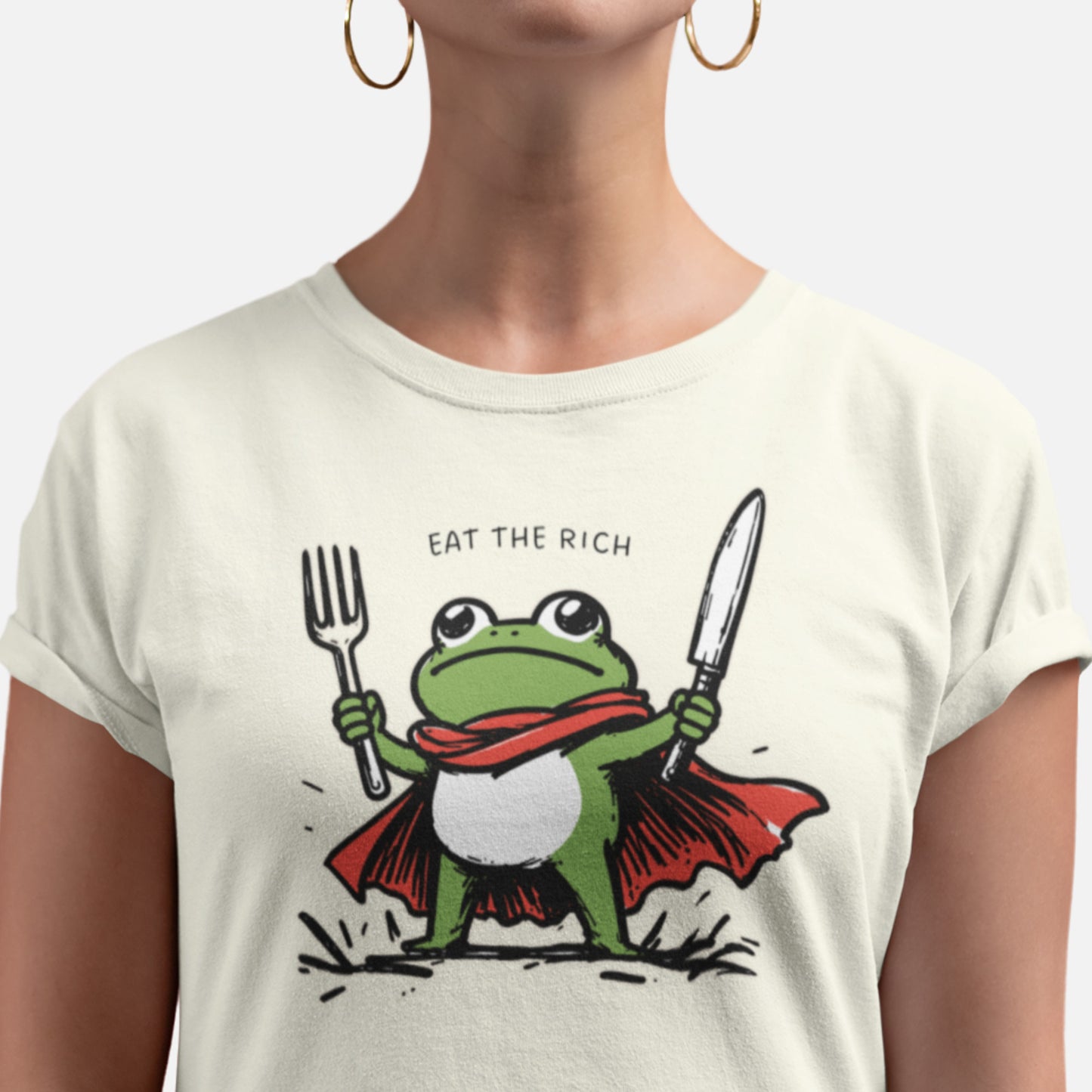 Person wearing a cream-colored unisex t-shirt featuring a cartoon frog hero holding a fork and knife beneath the phrase “Eat the Rich,” a humorous anti-corporate graphic combining political satire, protest art, and playful activist style.