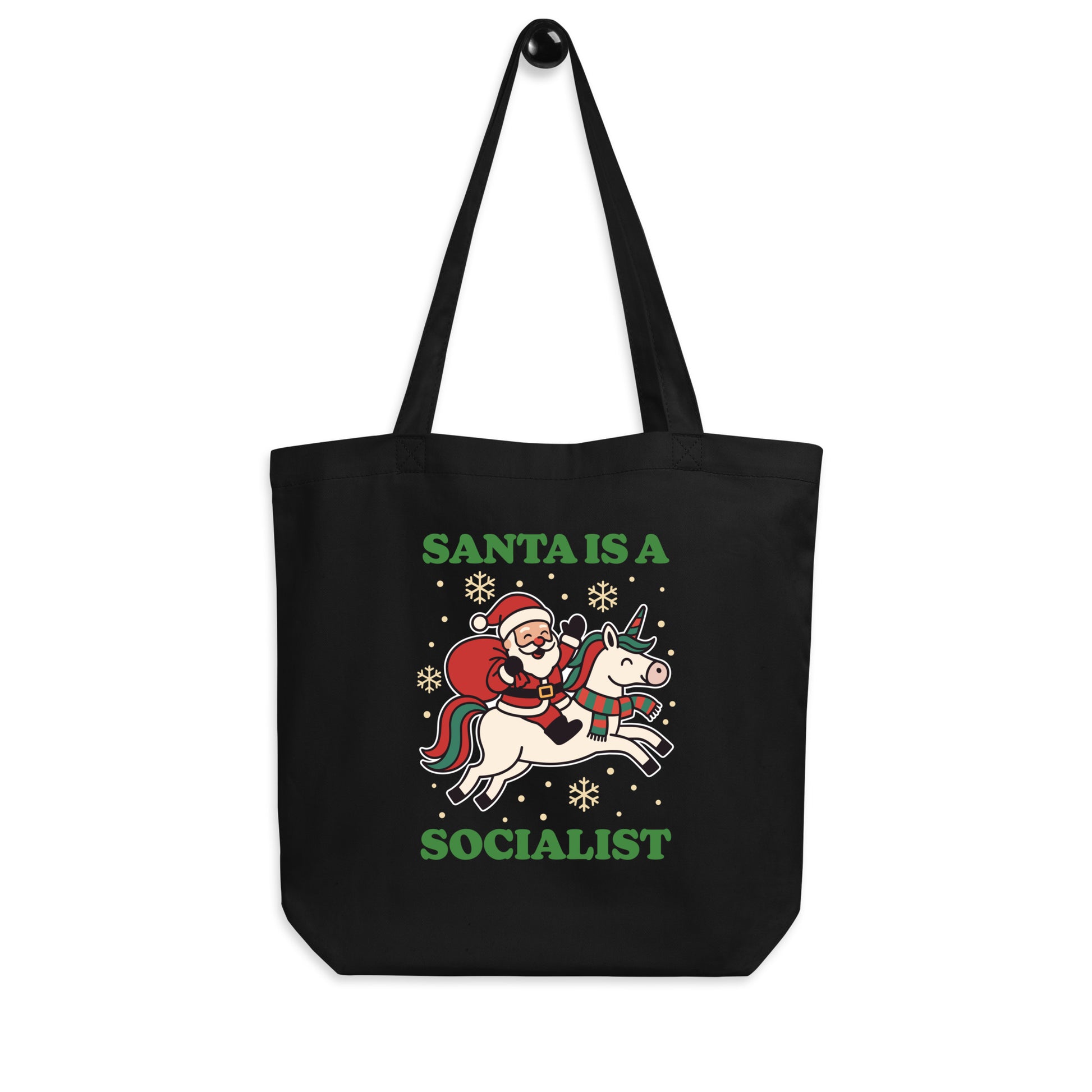 Black tote bag hanging on a hook, printed with a vintage-style Santa riding a unicorn illustration, holiday stars, and the message “SANTA IS A SOCIALIST,” from Blue Wave USA.