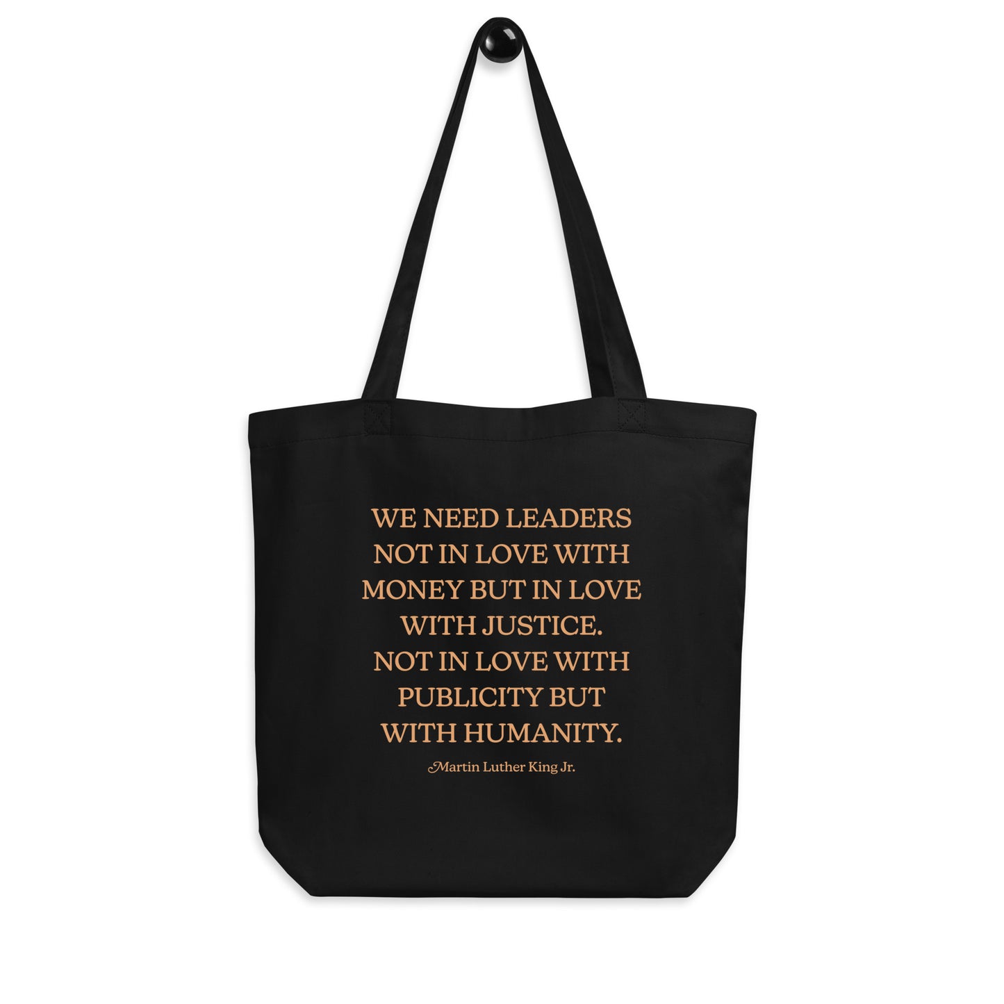 Black eco tote bag hanging on a hook, displaying the full MLK quote about justice, leadership, and humanity. A simple and bold Blue Wave USA activism design.