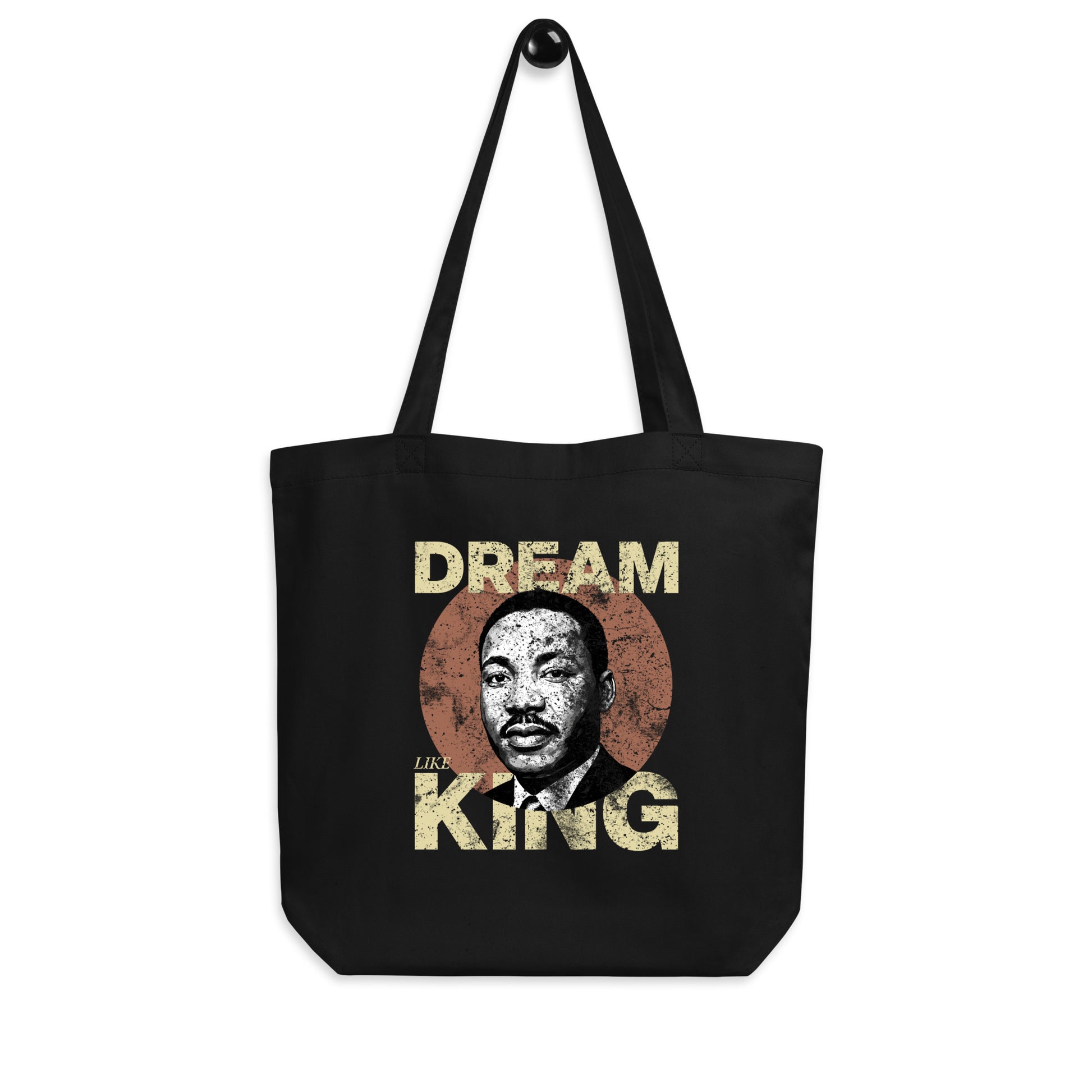Black eco tote bag hanging against a white backdrop, printed with the worn “DREAM LIKE KING” text and MLK portrait inside a rustic circle. Retro activist-style design.