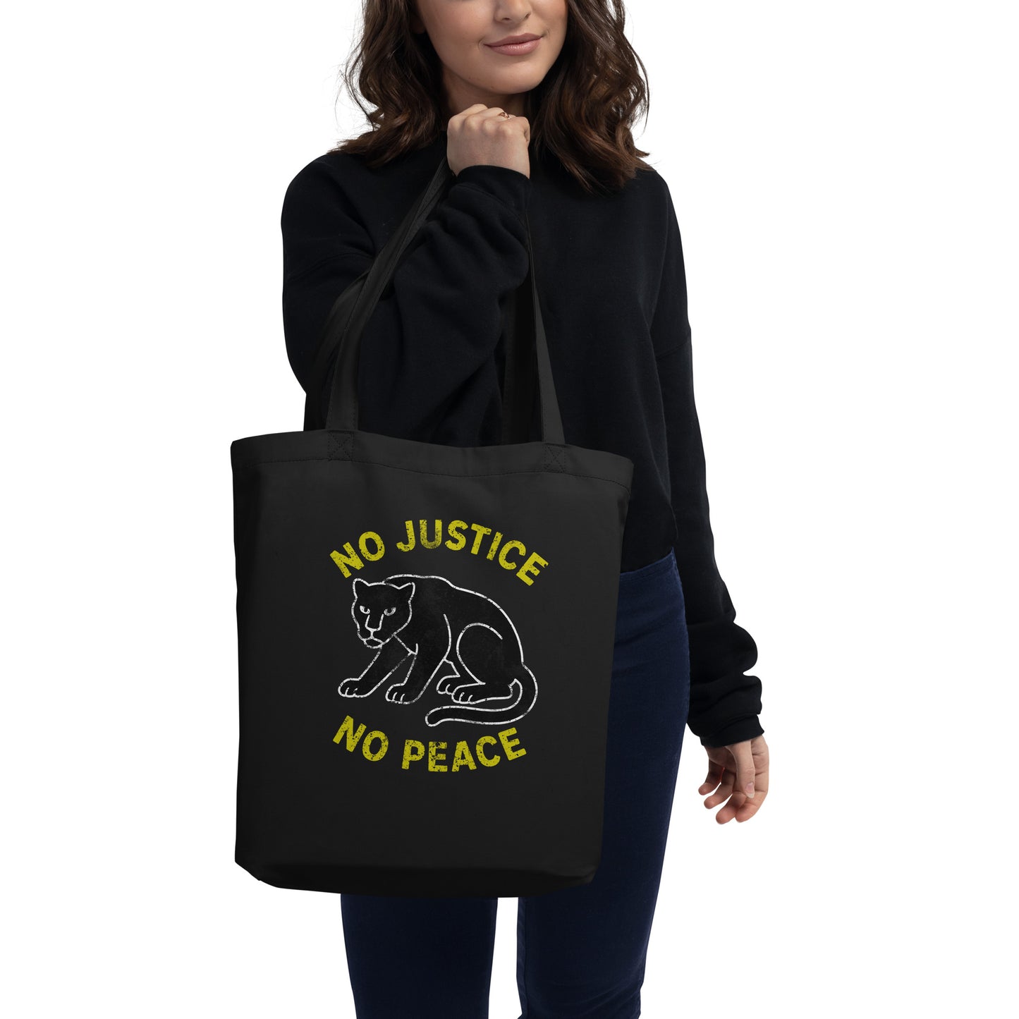 Person wearing dark sweater holding black eco tote bag with white panther artwork and yellow phrase “NO JUSTICE NO PEACE,” shown as a justice-focused design in everyday use.