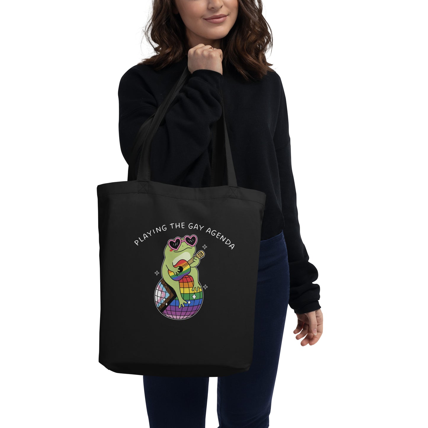Person carrying a black tote bag featuring a disco frog illustration, rainbow disco ball, and “Playing the Gay Agenda” design celebrating queer joy and self-expression.