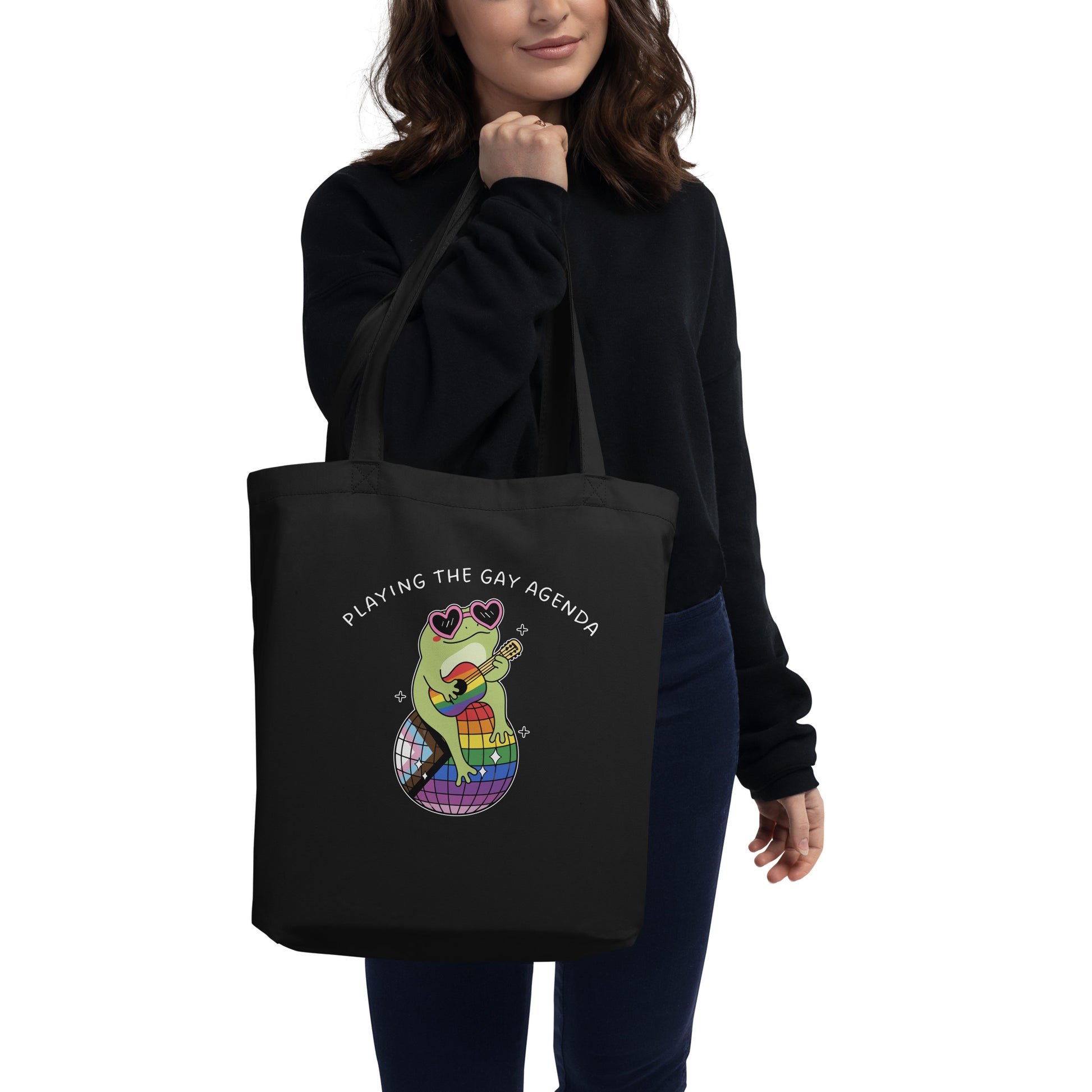 Person carrying a black tote bag featuring a disco frog illustration, rainbow disco ball, and “Playing the Gay Agenda” design celebrating queer joy and self-expression.