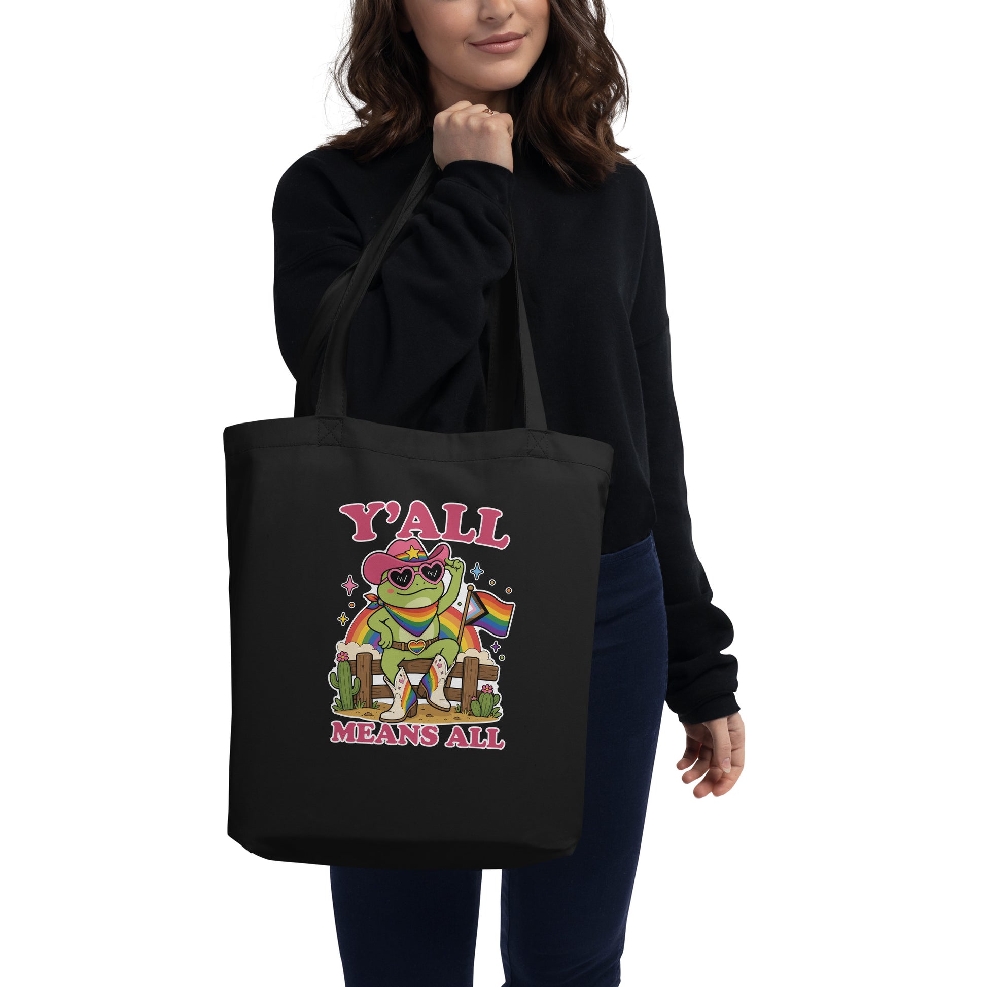 Person carrying a black eco tote bag featuring a rainbow cowboy frog holding a pride flag with cheerful “Y’All Means All” message.
