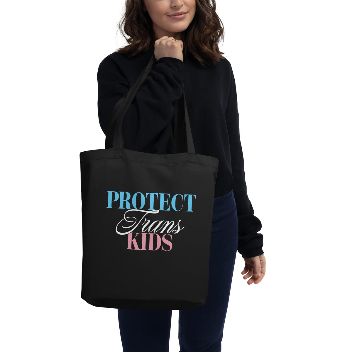 Person carrying a black eco tote bag featuring the “Protect Trans Kids” design in trans pride-inspired blue, white, and pink lettering.
