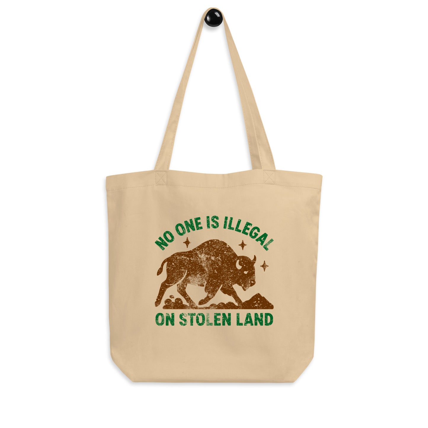 Beige tote bag hanging from a hook, showcasing the buffalo silhouette and the phrase “NO ONE IS ILLEGAL ON STOLEN LAND,” styled in warm, earthy activist colors.