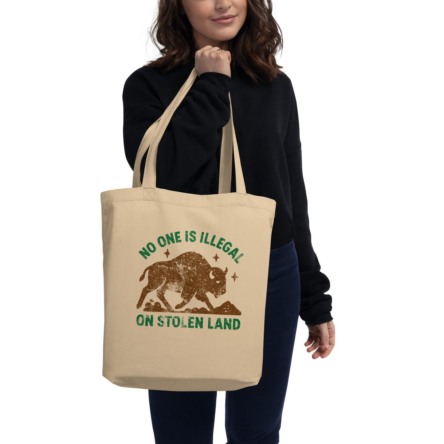 Model holding a beige canvas tote printed with a distressed buffalo graphic and the message “NO ONE IS ILLEGAL ON STOLEN LAND,” a casual anti-colonial accessory from Blue Wave USA.