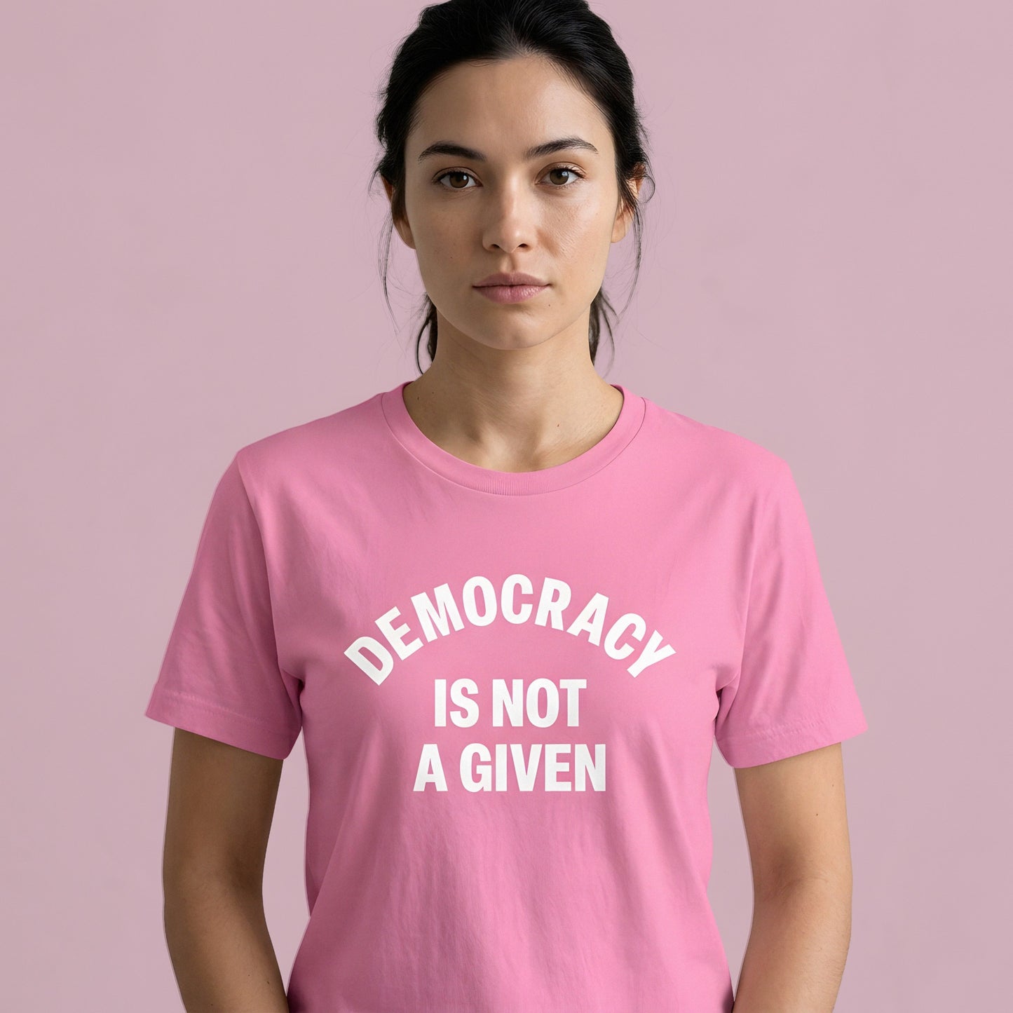 Woman wearing pink political t-shirt featuring bold white “Democracy Is Not a Given” typography against soft pink studio background.