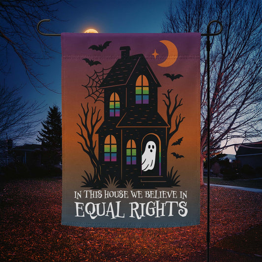 “In This House We Believe in Equal Rights” Halloween garden flag with haunted house, ghost, bats, and crescent moon, displayed outdoors at night.
