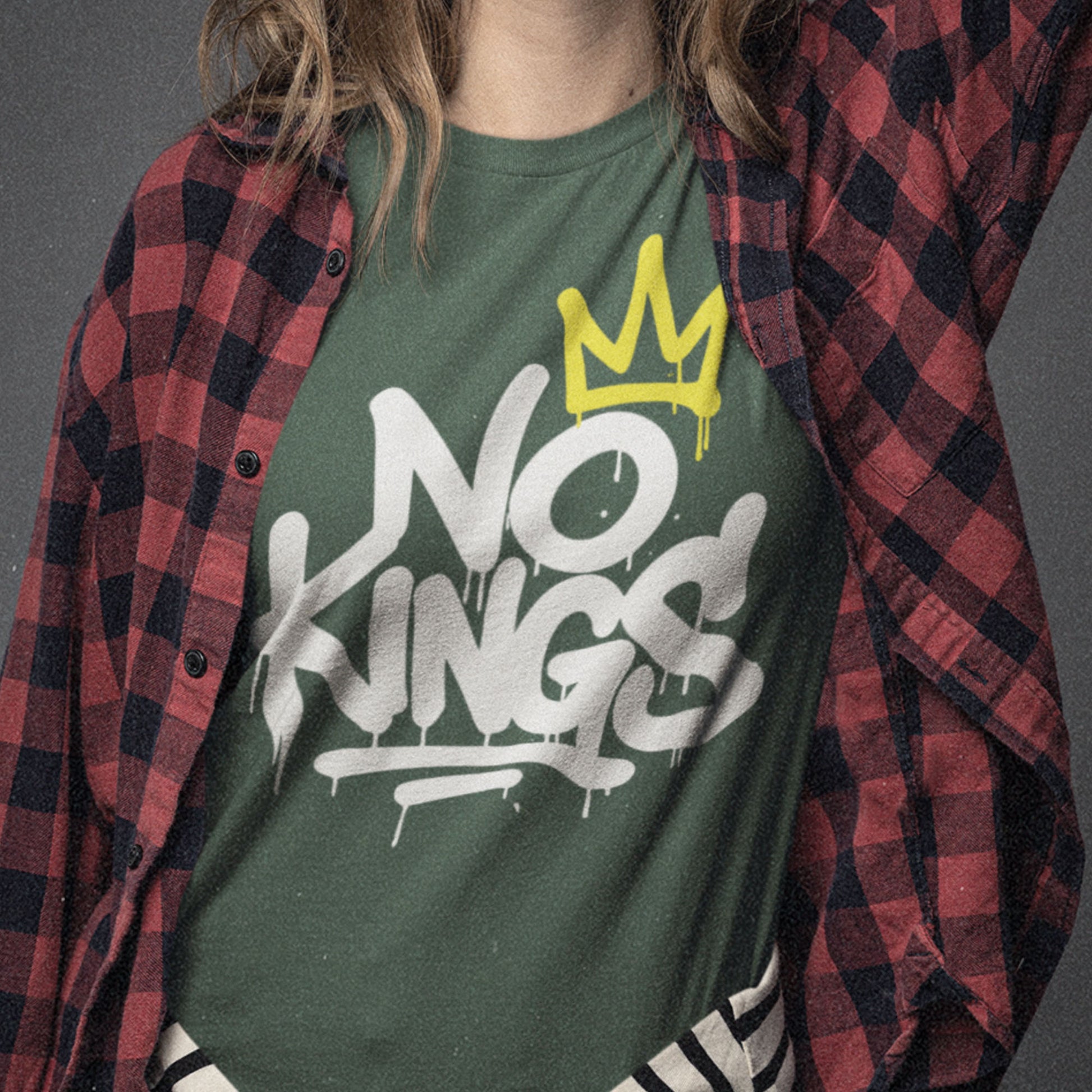 Close-up of a person wearing a forest green “No Kings” t-shirt with bold white graffiti lettering and a yellow crown graphic. The shirt is layered under an open red-and-black plaid flannel, giving a relaxed streetwear look.