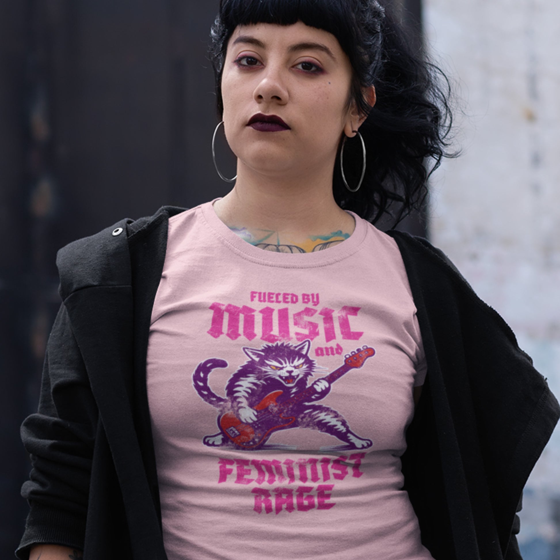 Model wearing a light pink unisex t-shirt with a punk rock guitar-playing cat illustration and the phrase “FUELED BY MUSIC AND FEMINIST RAGE,” styled outdoors from Blue Wave USA.