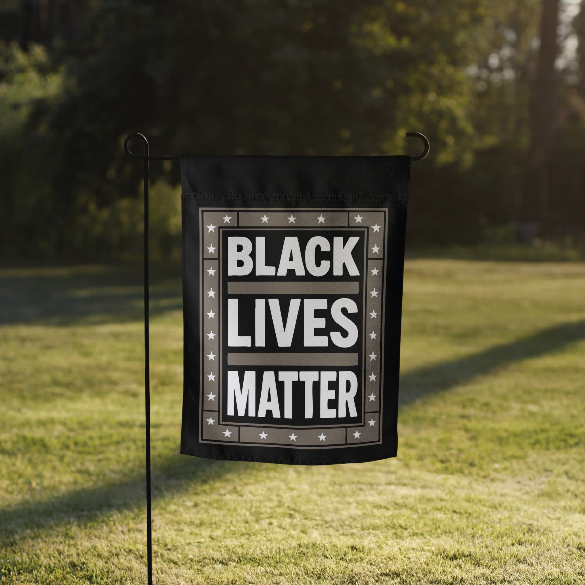 Black Lives Matter garden flag with star border shown on a flag stand in a grassy yard with sunlight behind it.