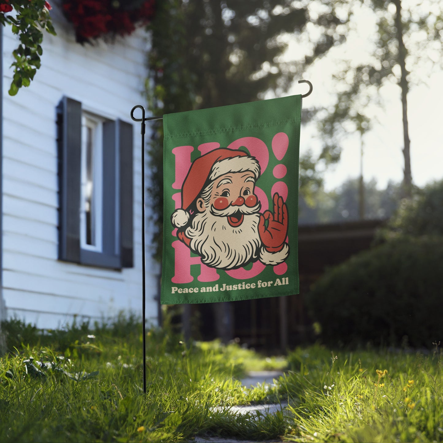 Green garden flag displayed on a metal stand outside a home, showing a vintage Santa graphic, pink “HO!” lettering, and the message “Peace and Justice for All.”