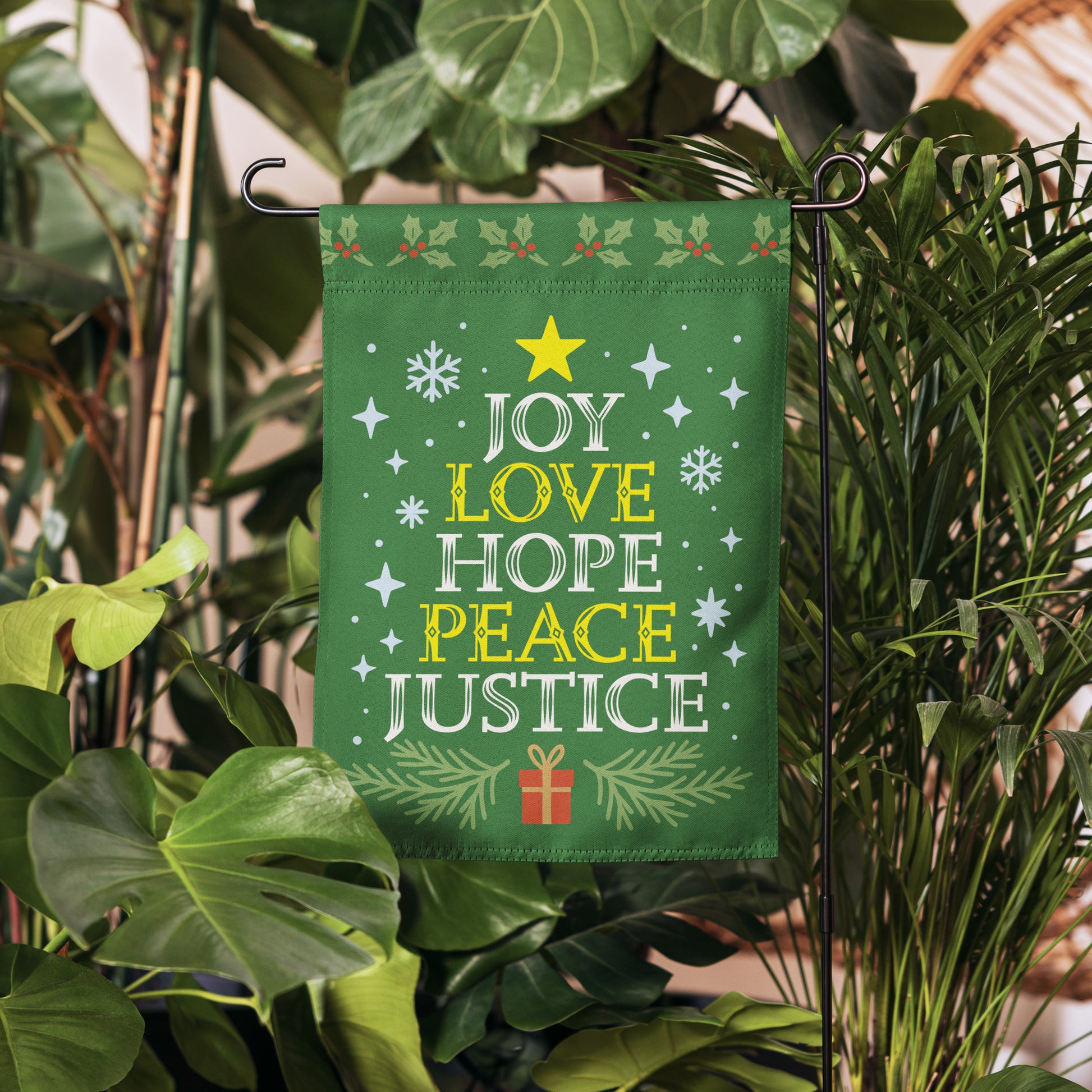 Green garden flag displayed in a garden setting, featuring decorative stars, snowflakes, greenery, and the printed message “JOY LOVE HOPE PEACE JUSTICE.”