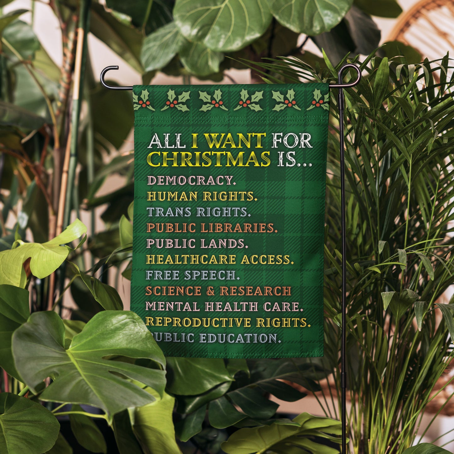 Green plaid garden flag hanging in a garden setting, featuring holly accents and the printed list “ALL I WANT FOR CHRISTMAS IS…” with items like democracy, human rights, and public libraries.