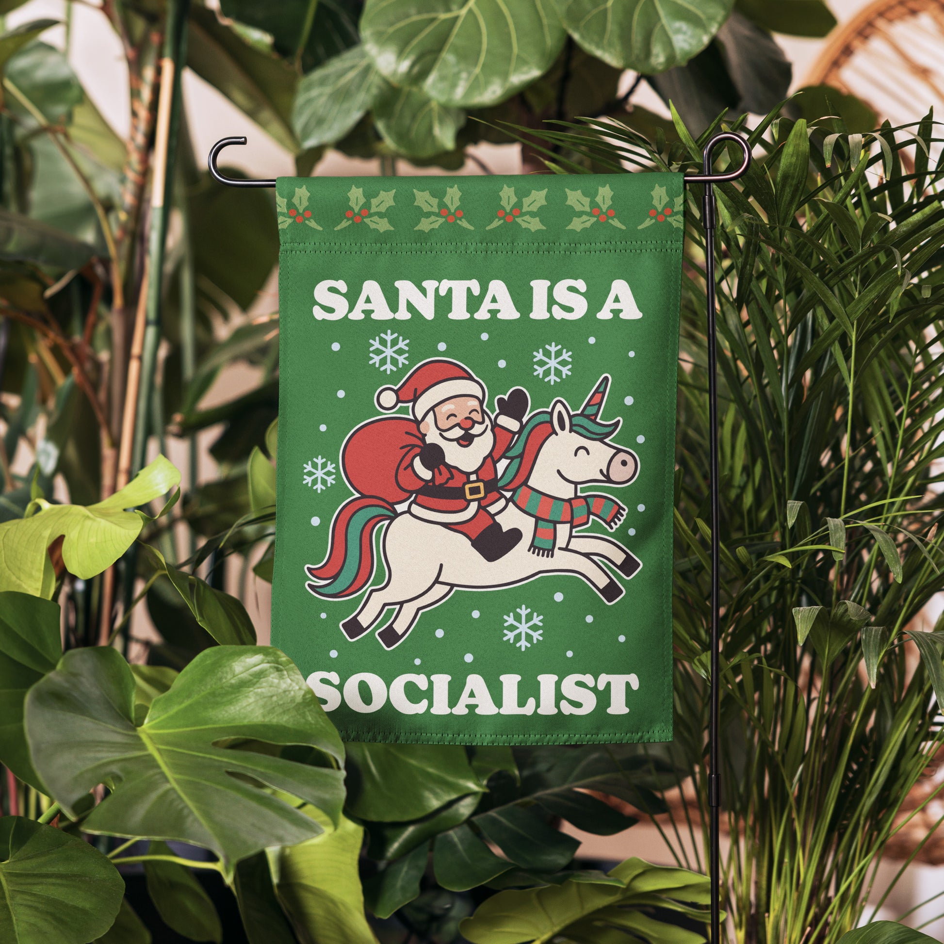 Green garden flag hanging in a garden setting, printed with a vintage Santa riding a unicorn, white snowflakes, and the message “SANTA IS A SOCIALIST.”
