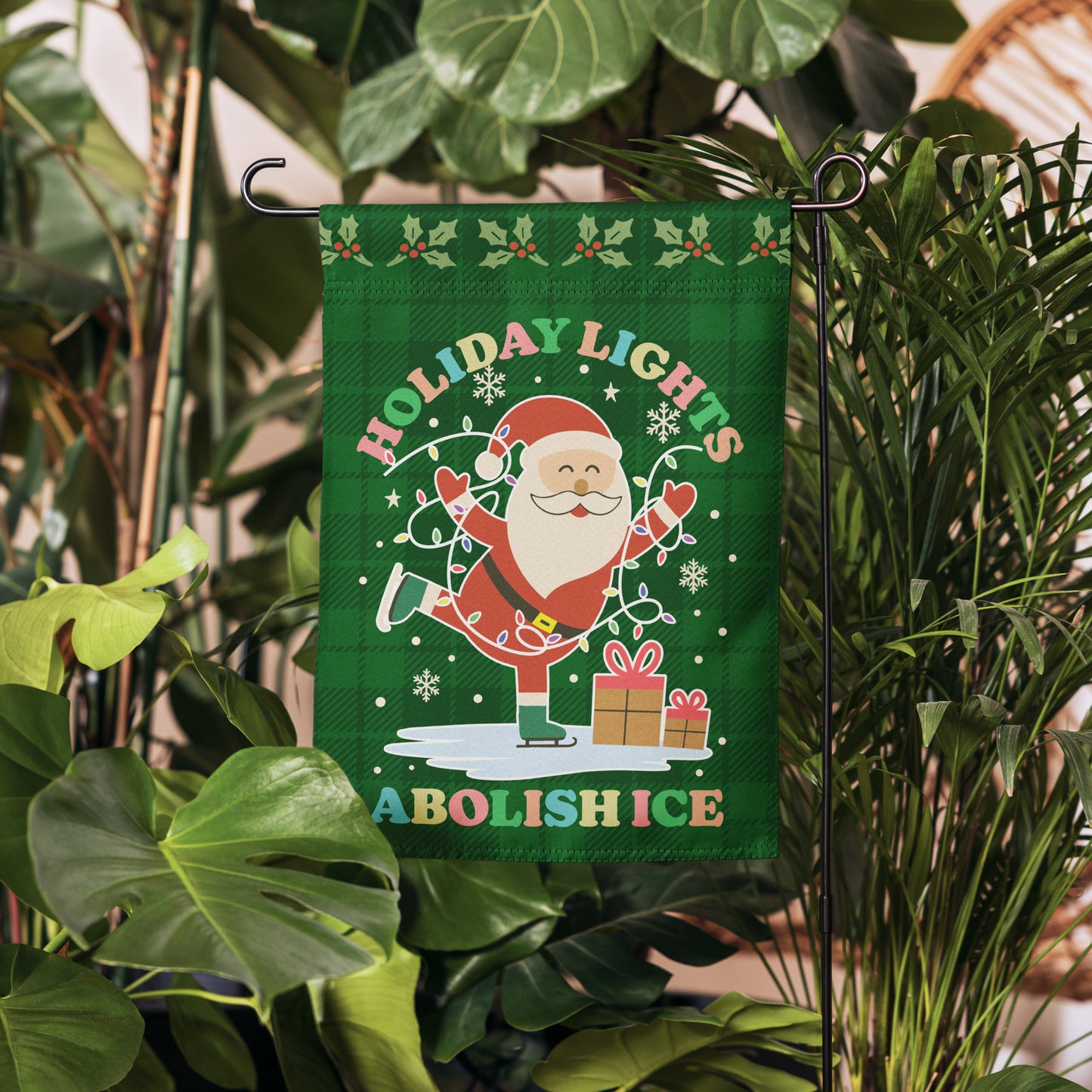 A green garden flag showing Santa, colorful lights, stars, and presents arranged around the printed message “HOLIDAY LIGHTS ABOLISH ICE,” hanging among lush plants for cheerful activist holiday style from Blue Wave USA.