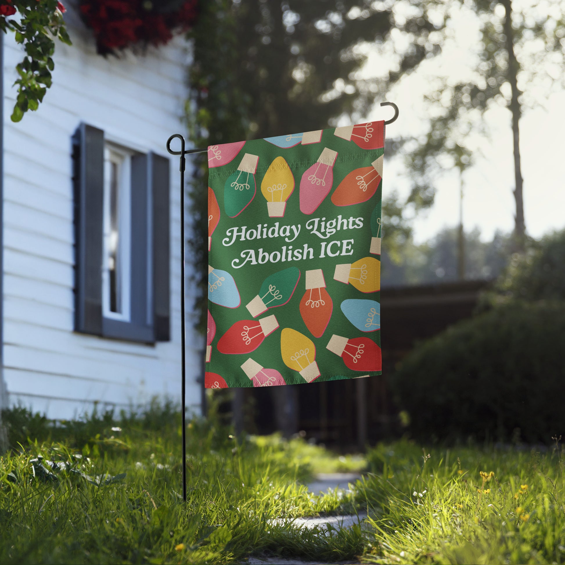 A green garden flag featuring festive multicolor bulbs framing the message “Holiday Lights Abolish ICE,” shown in a sunny yard setting for progressive holiday décor from Blue Wave USA.