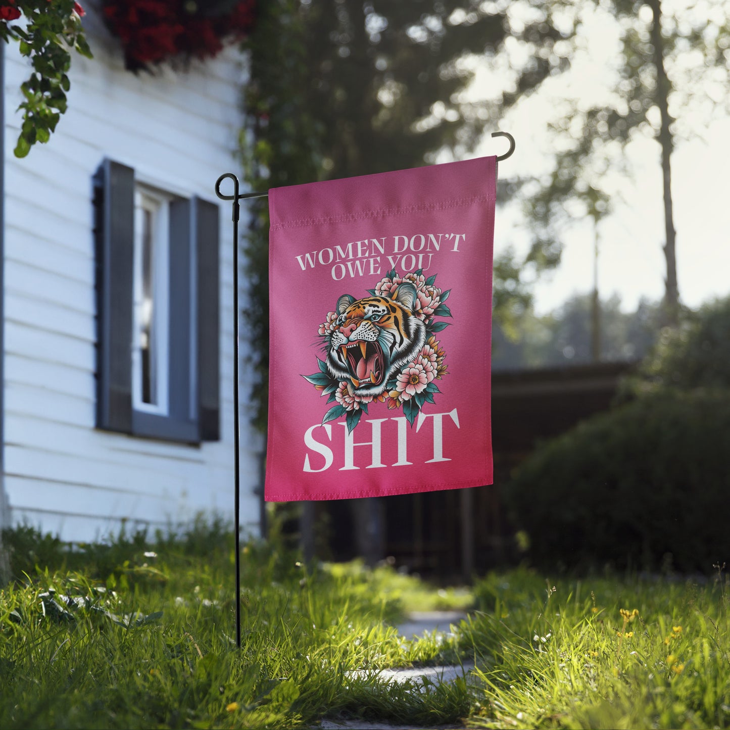 Garden flag planted near a house and lawn, showcasing the illustrated tiger surrounded by flowers with strong feminist text reading “WOMEN DON’T OWE YOU SH*T,” from Blue Wave USA.