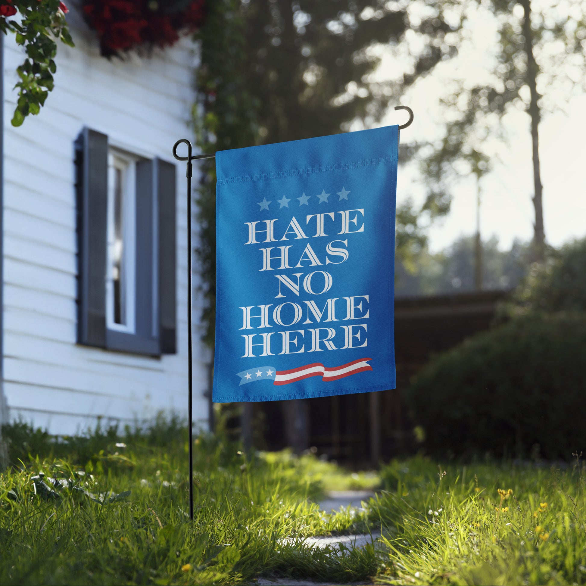 Garden flag mounted on metal stand in grassy yard, blue background with white text reading “HATE HAS NO HOME HERE,” visible from sidewalk view.