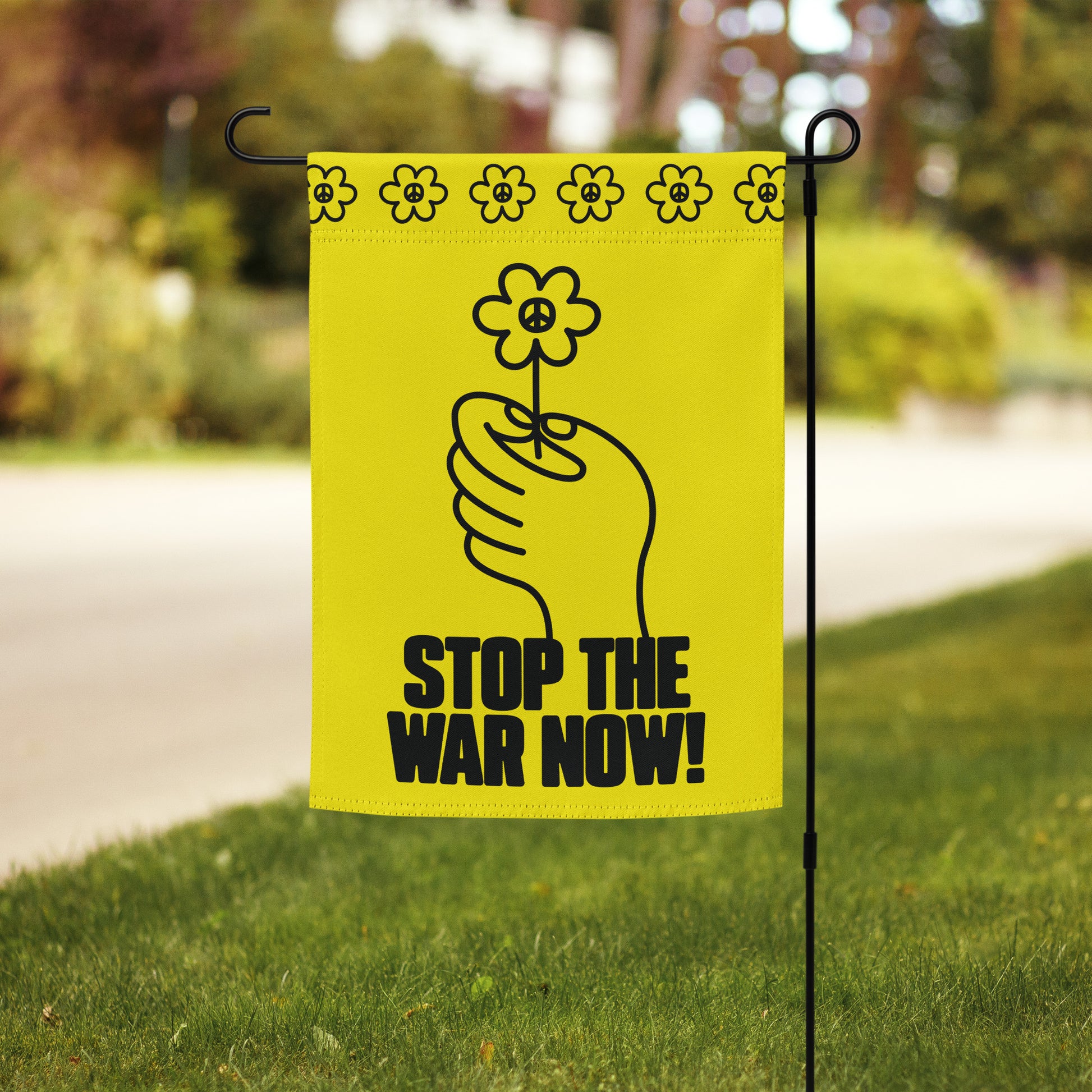 Yellow garden flag with hand-and-flower illustration and “STOP THE WAR NOW!” protest wording hanging beside suburban walkway.
