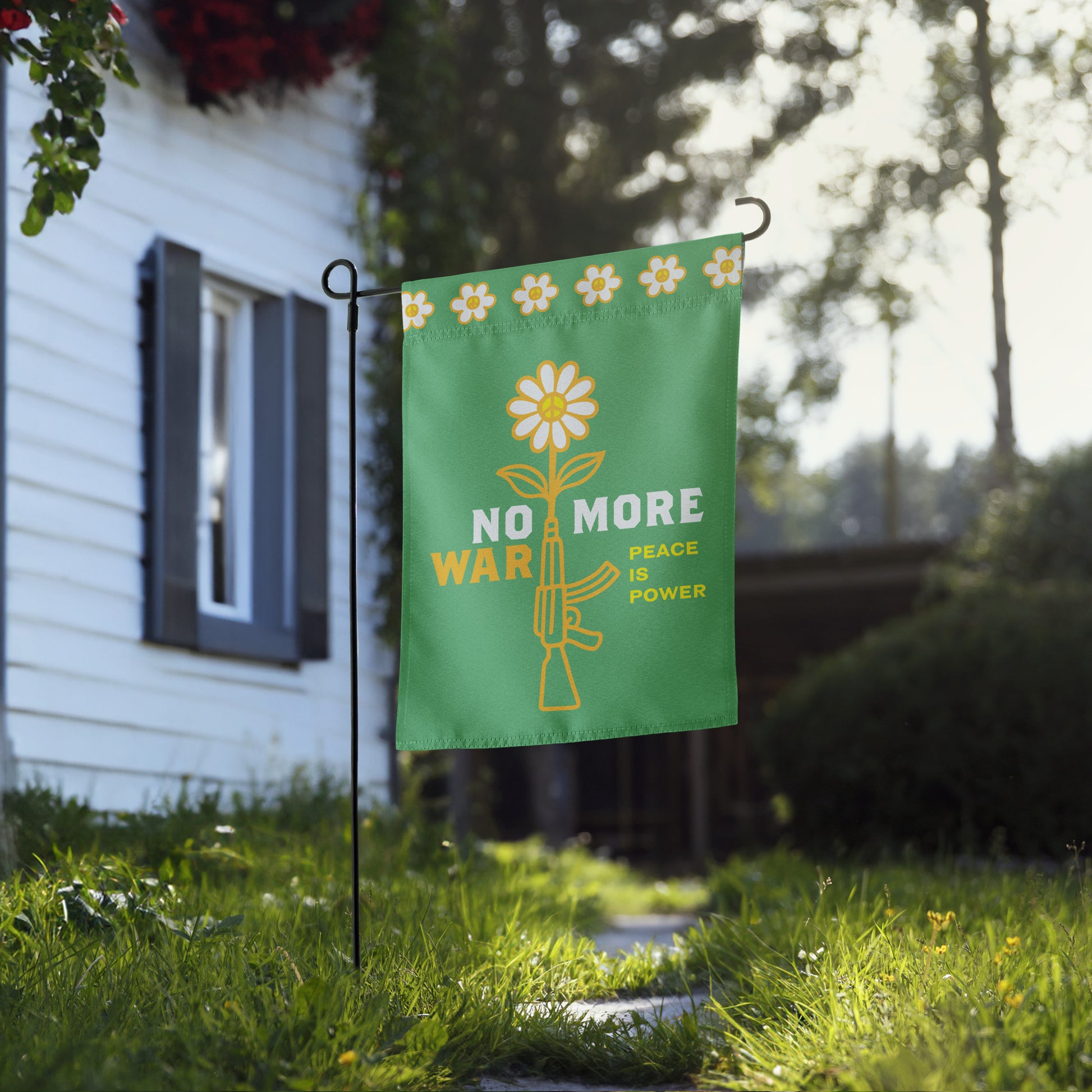 Green garden flag with floral rifle illustration and “NO MORE WAR” peace statement hanging beside home walkway in yard setting.
