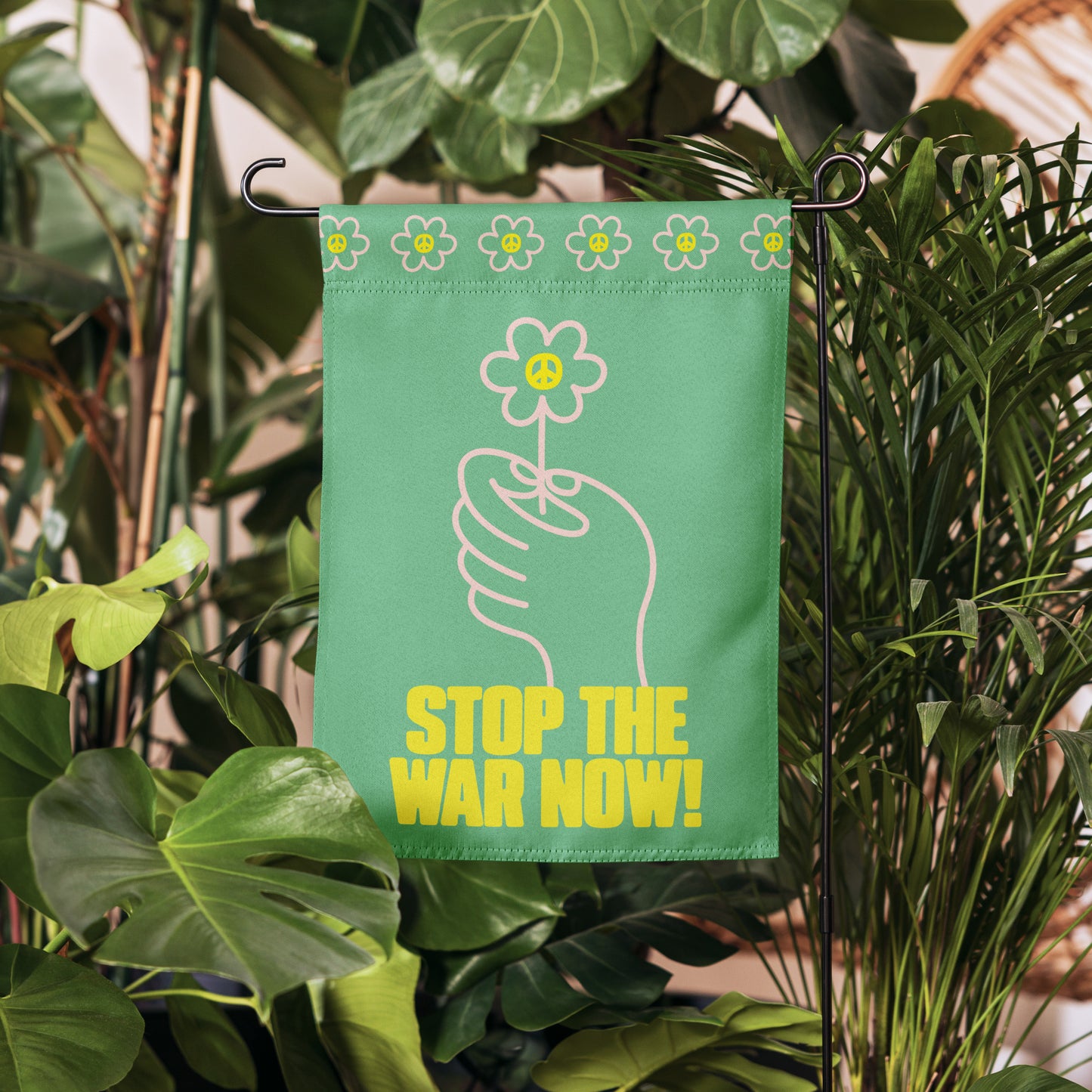 Anti-war garden flag with floral hand graphic and “STOP THE WAR NOW!” wording hanging near leafy plants in outdoor walkway scene.