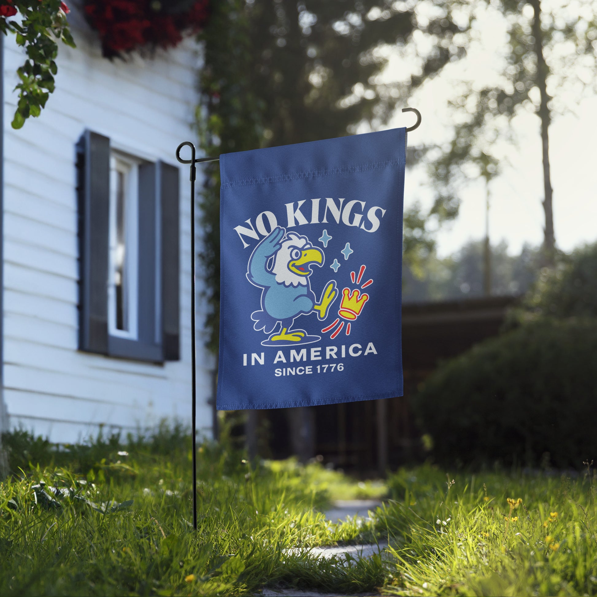 Blue garden flag hanging near home walkway showing eagle illustration and text reading “NO KINGS IN AMERICA SINCE 1776.”