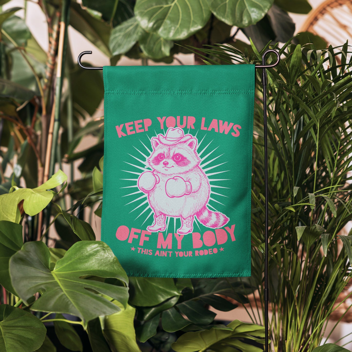 Blue Wave USA garden flag displayed among greenery, showcasing raccoon cowboy illustration and reproductive rights message in justice-focused yard decor styling.