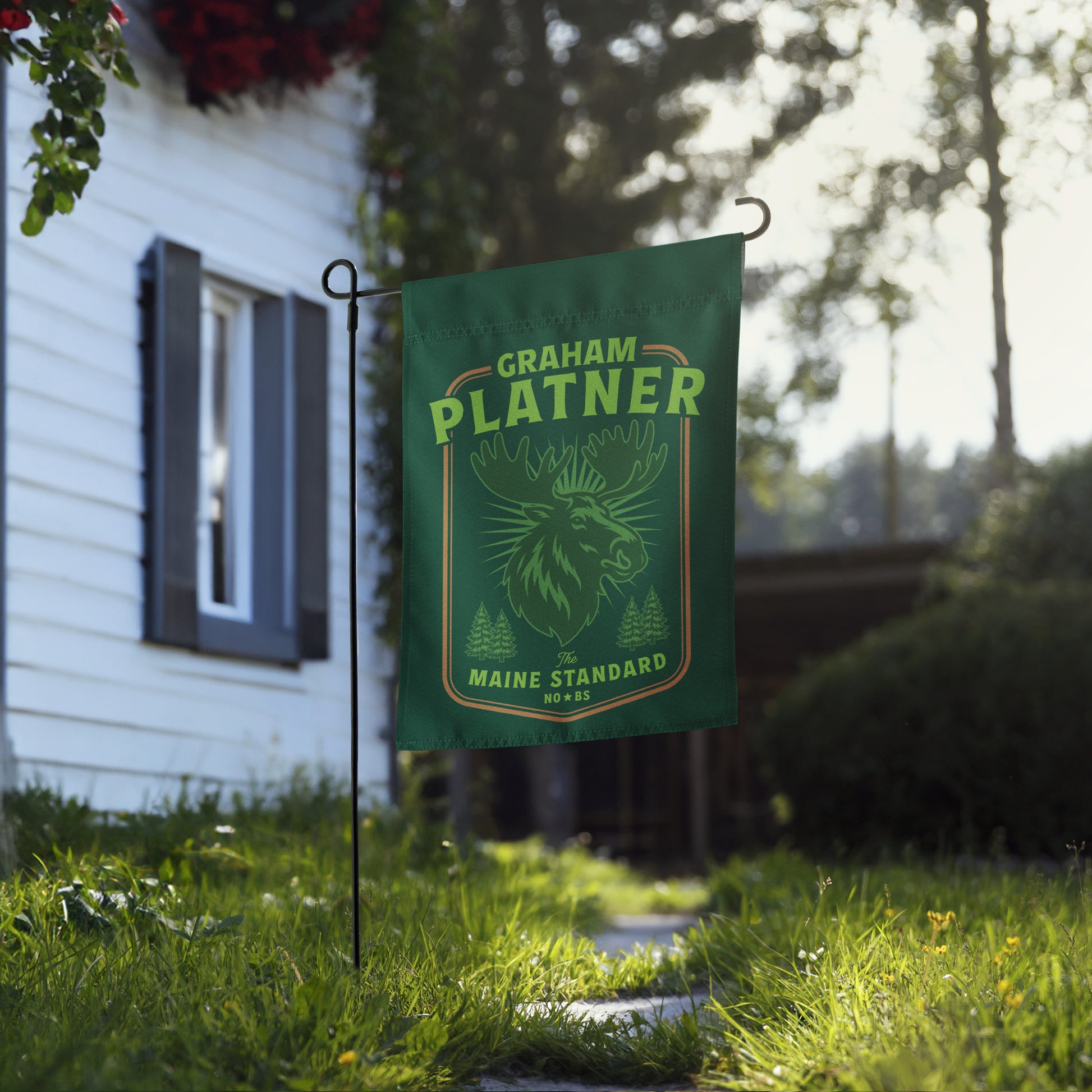 Garden flag featuring green moose illustration and “Graham Platner Maine Standard” text shown near house walkway in natural outdoor setting.