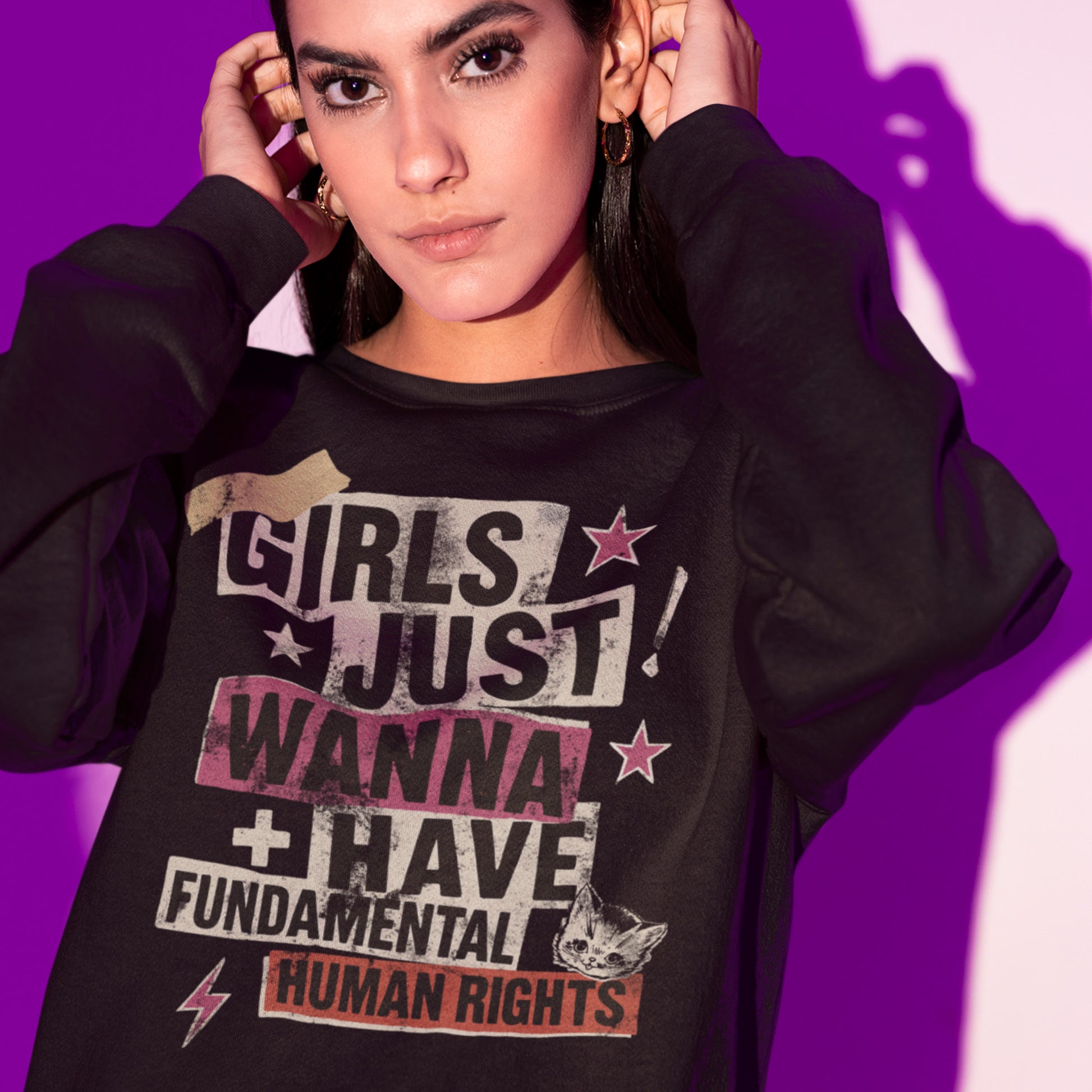 Model wearing a black sweatshirt that reads “Girls Just Wanna Have Fundamental Human Rights” in bold punk collage lettering with taped textures, stars, and a lightning bolt. The design stands out in pink, beige, and gray tones against a bright purple background.