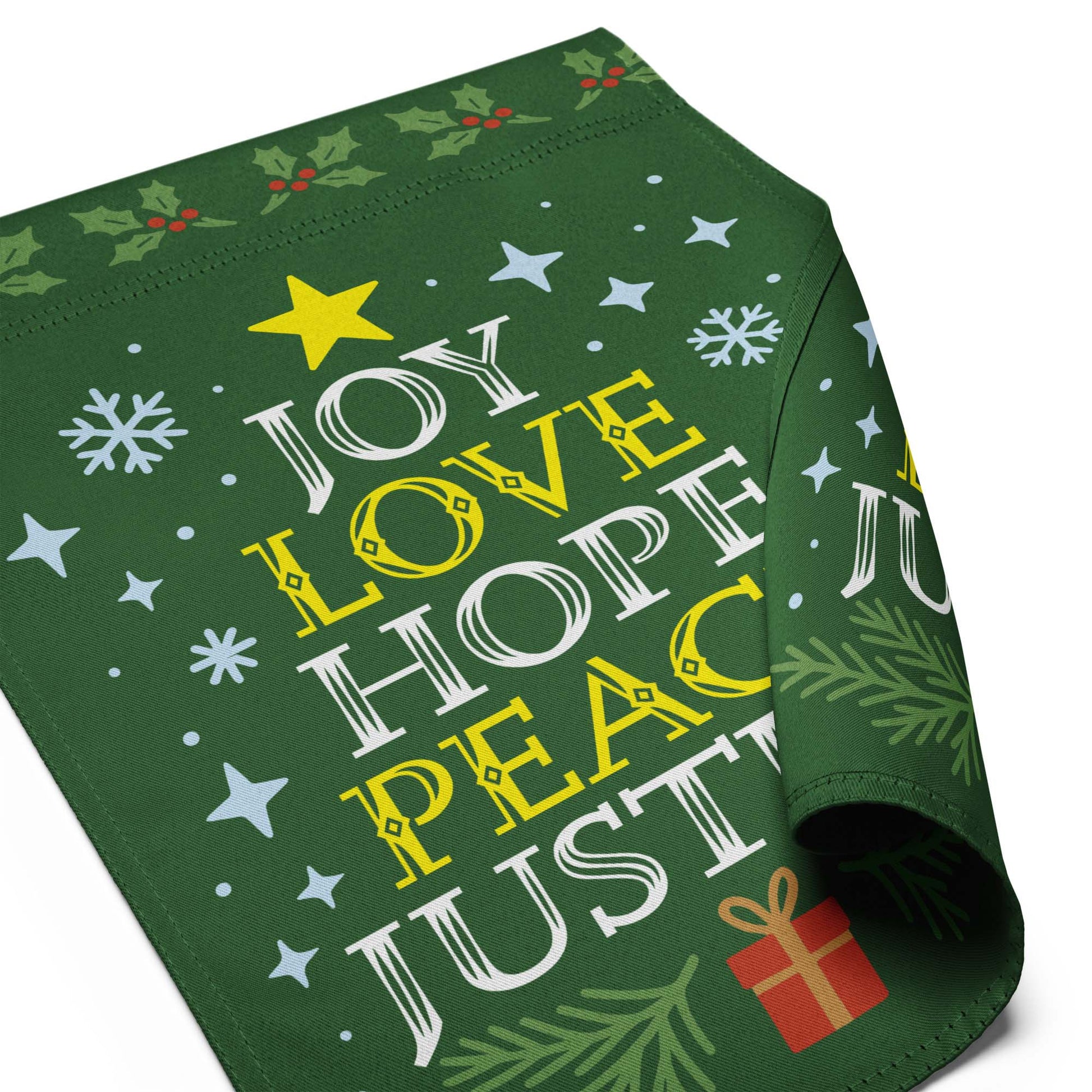 Close-up of a folded green garden flag displaying part of the printed message “JOY LOVE HOPE PEACE JUSTICE” along with snowflakes and greenery.