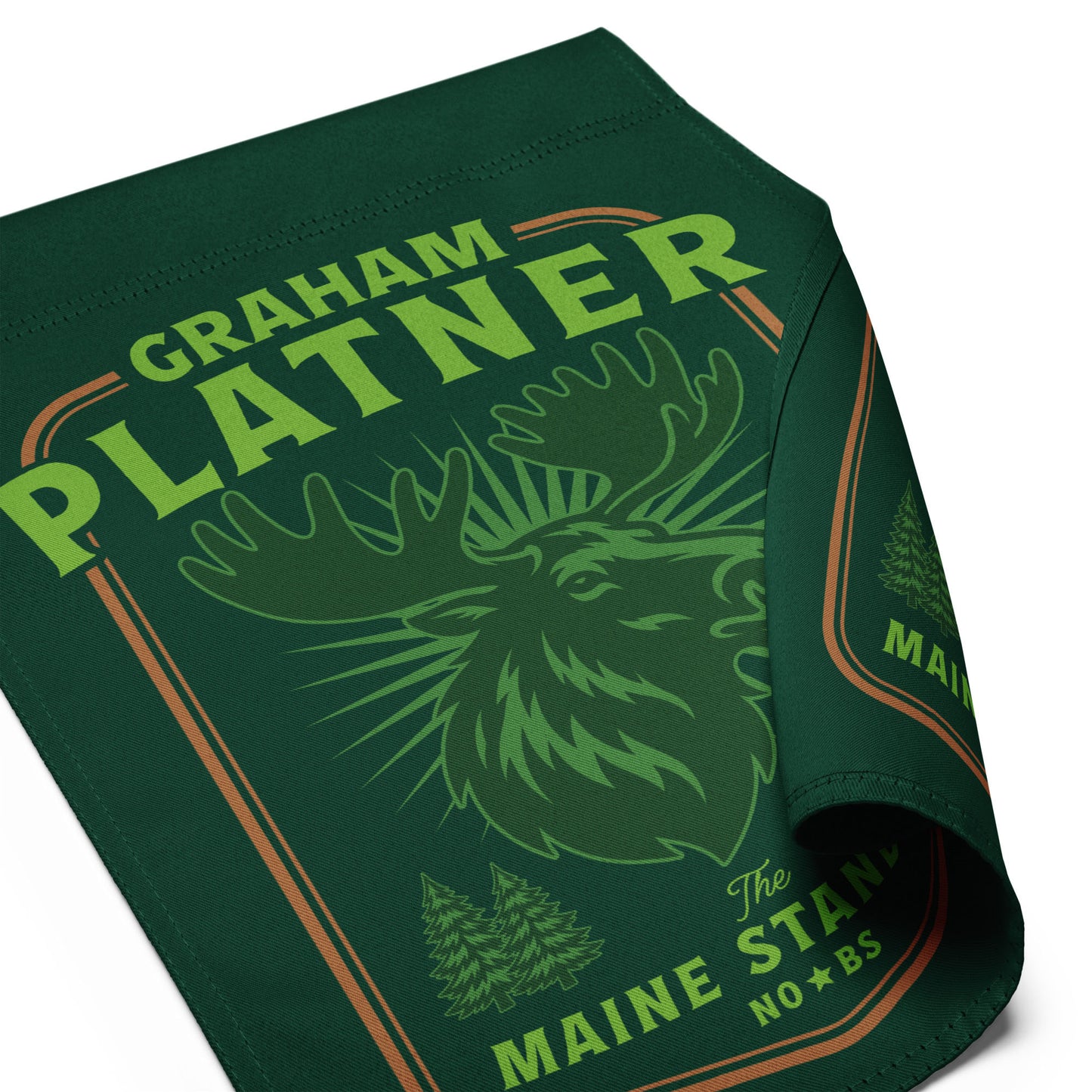 Close-up of green garden flag fabric showing moose illustration and “Graham Platner Maine Standard” text with detailed vintage outdoor print.