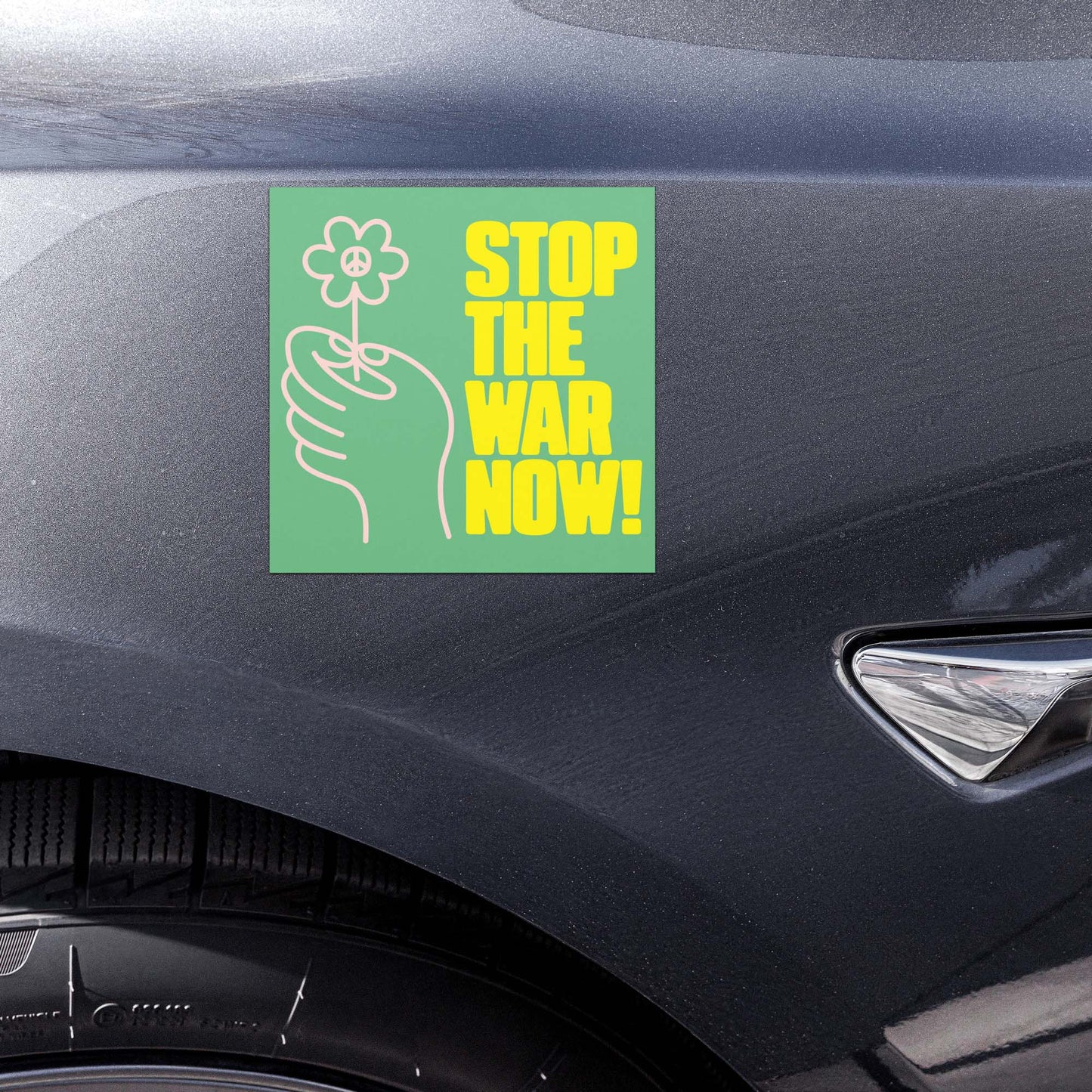 Green anti-war car magnet with floral hand graphic and “STOP THE WAR NOW!” protest wording shown on vehicle door panel.
