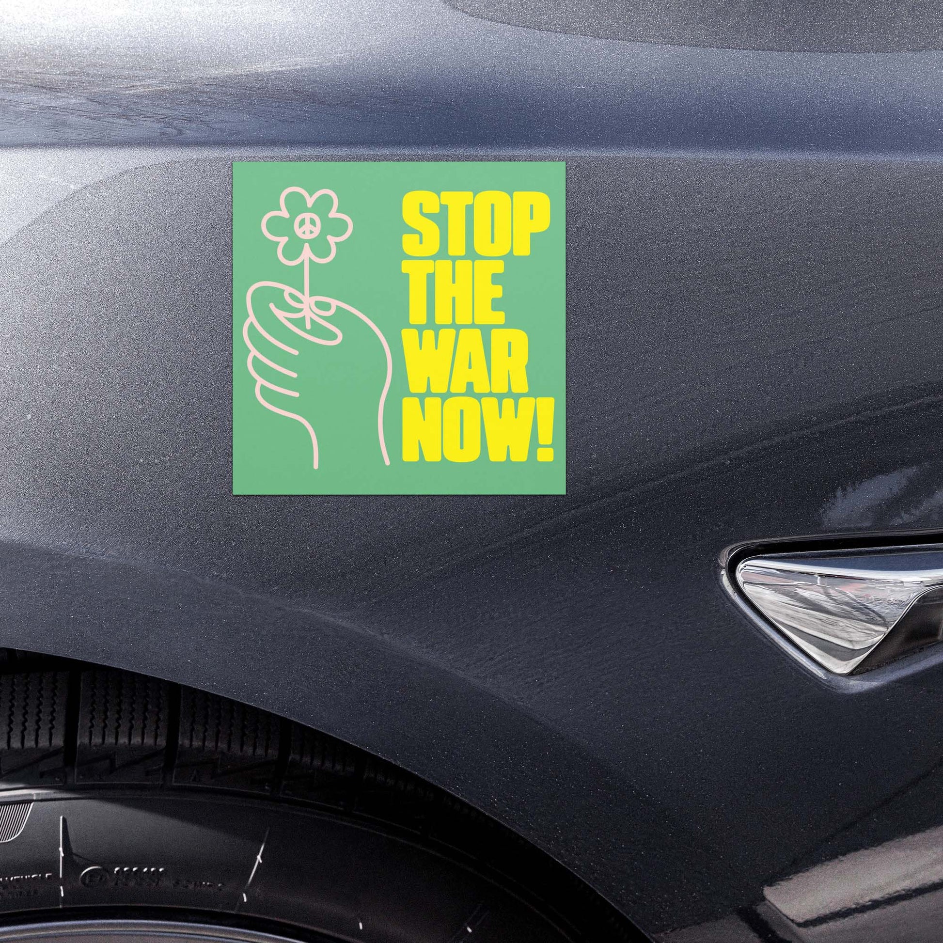 Green anti-war car magnet with floral hand graphic and “STOP THE WAR NOW!” protest wording shown on vehicle door panel.