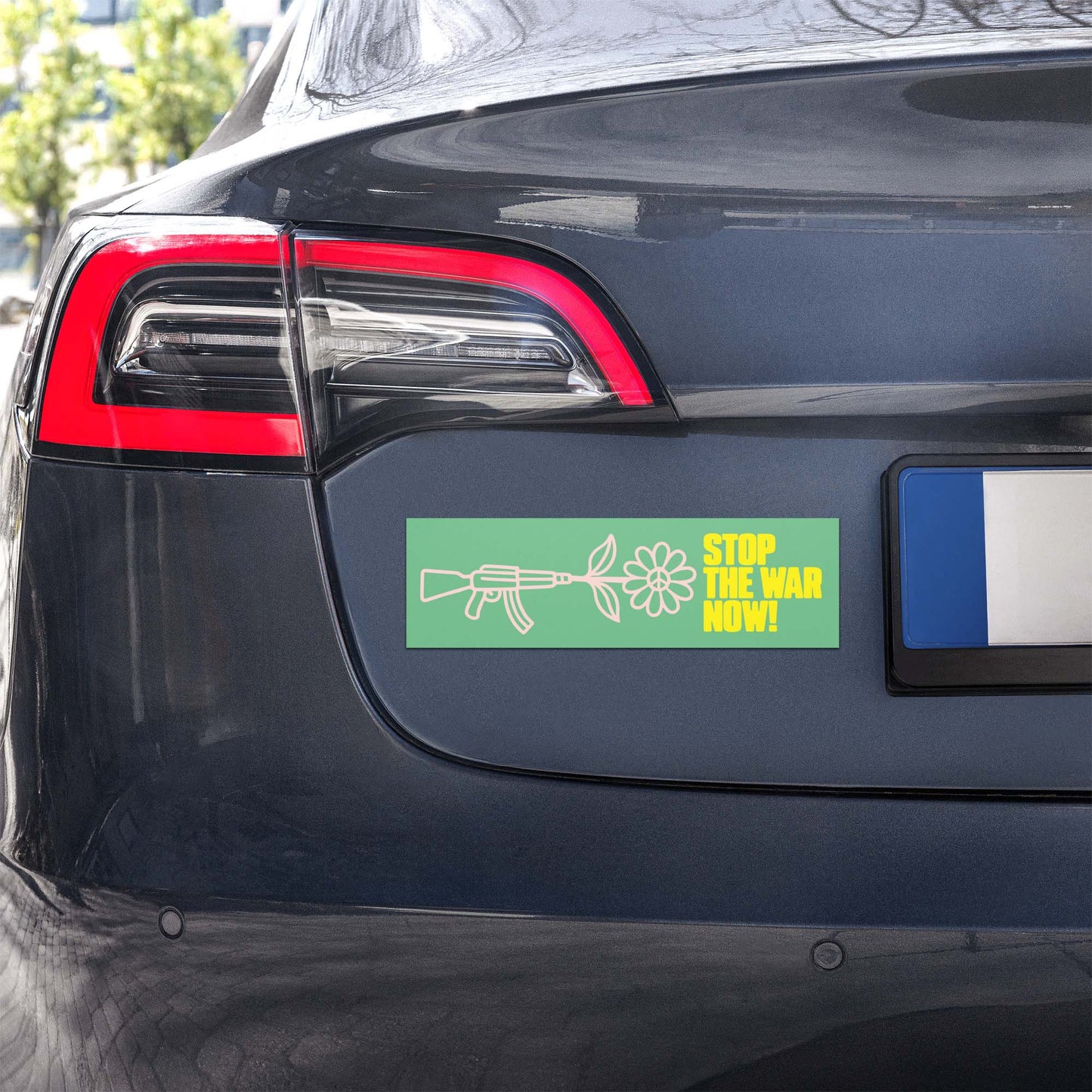 Green bumper magnet with floral rifle illustration and “STOP THE WAR NOW!” protest wording shown on rear of vehicle.