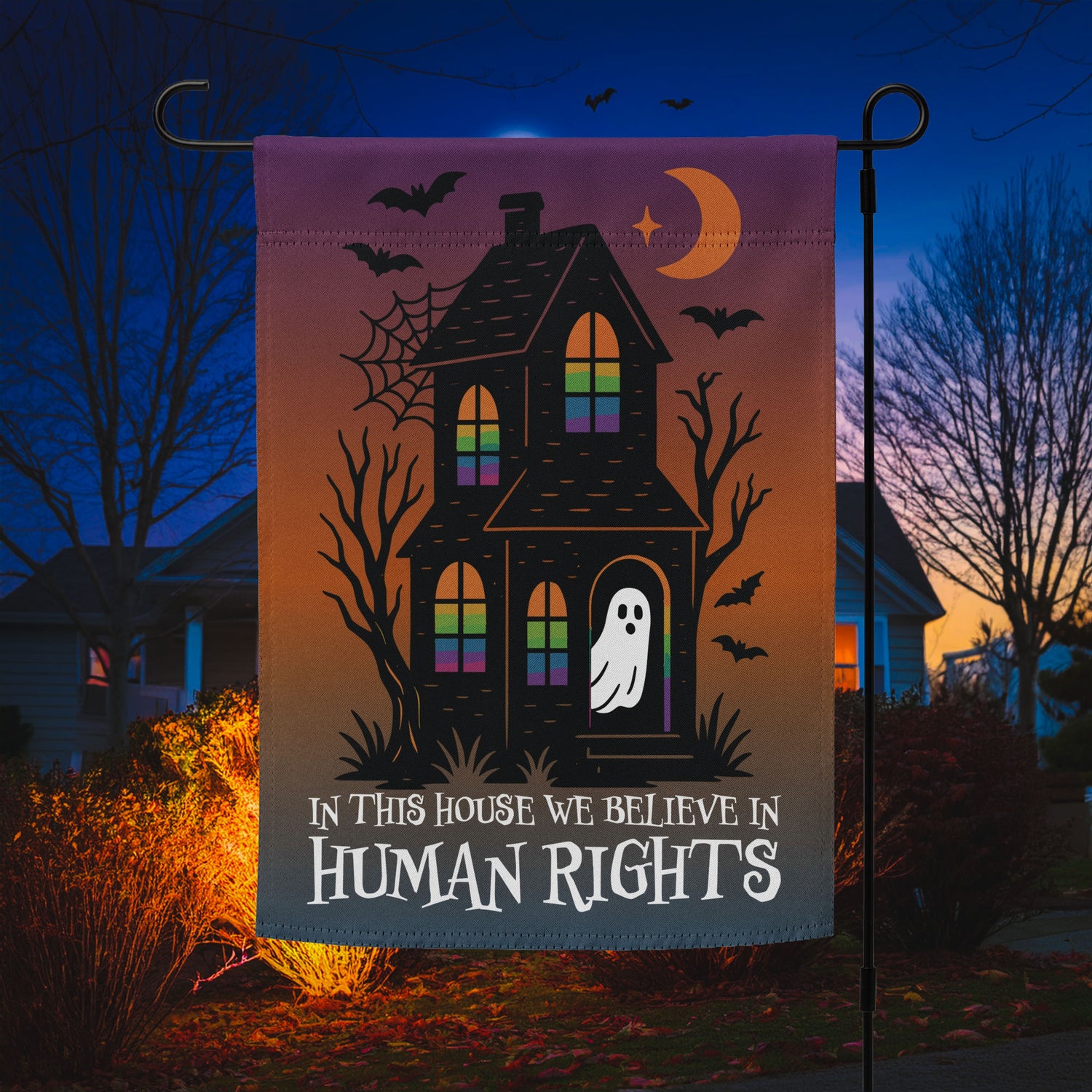 “In This House We Believe in Human Rights” Halloween garden flag featuring a haunted house, bats, and ghost, displayed outdoors at night.