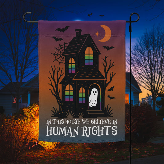 “In This House We Believe in Human Rights” Halloween garden flag featuring a haunted house, bats, and ghost, displayed outdoors at night.
