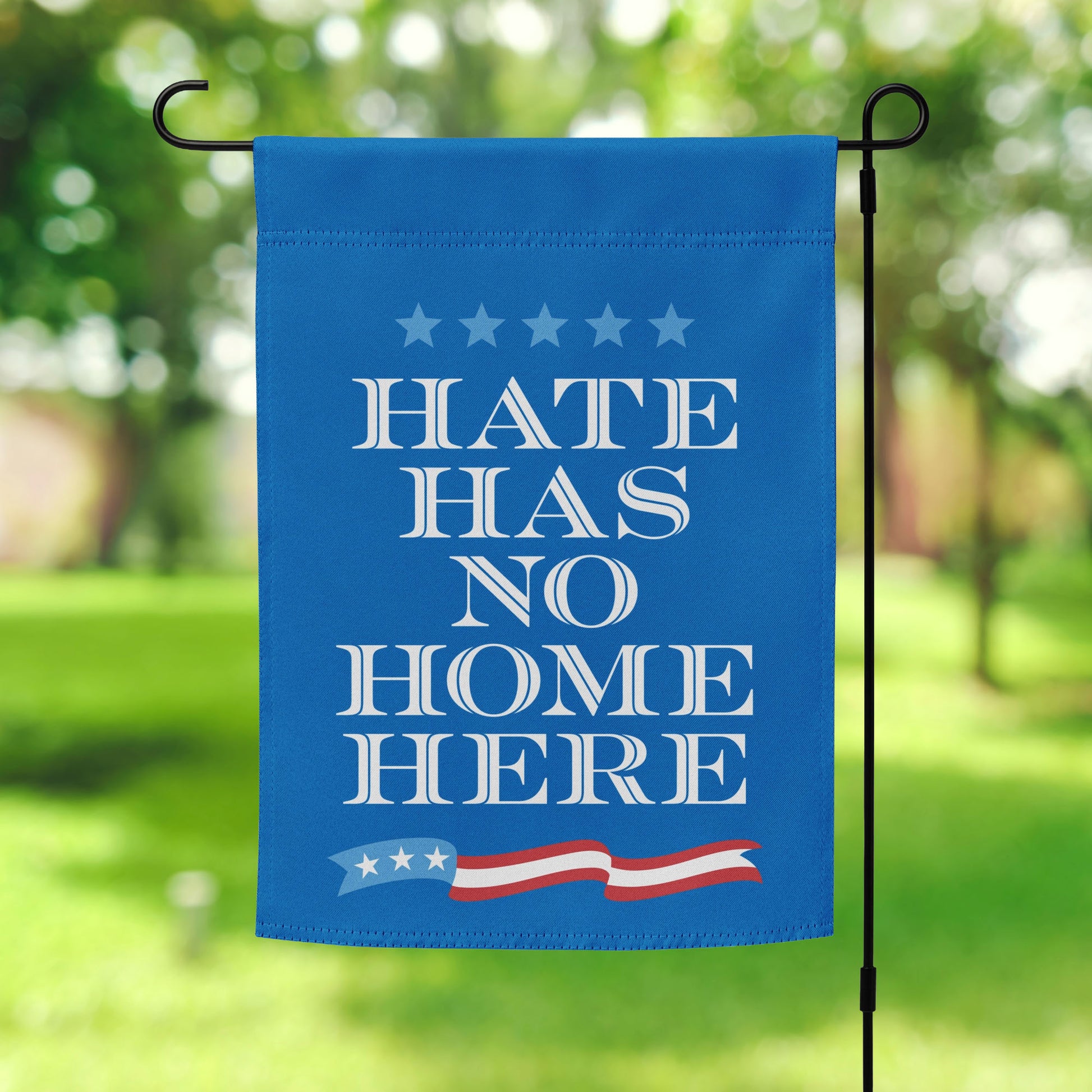 Blue garden flag featuring stacked white serif text reading “HATE HAS NO HOME HERE,” star accents above and red-white stripe motif below, vertical layout.