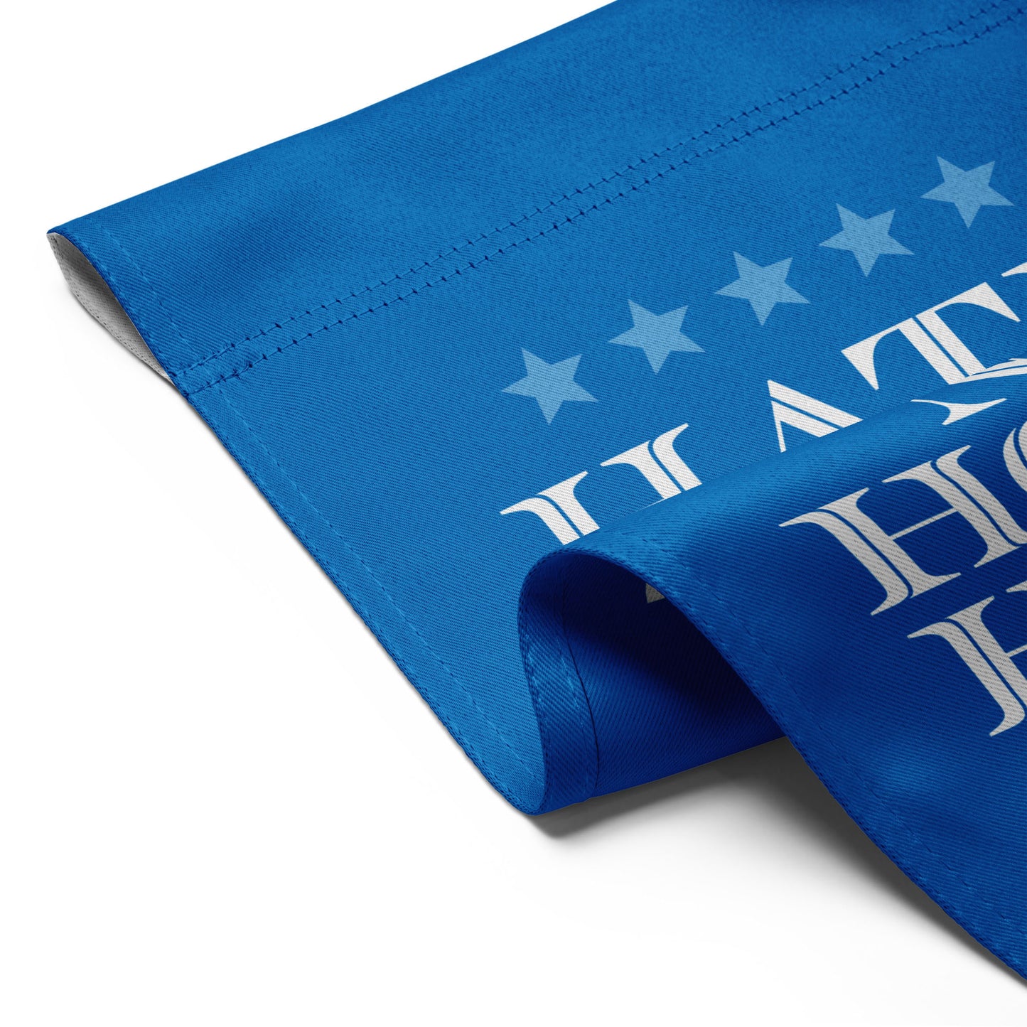 Close-up of blue garden flag fabric showing stitched edges, white serif text “HATE HAS NO HOME HERE,” star accents and red stripe detail.