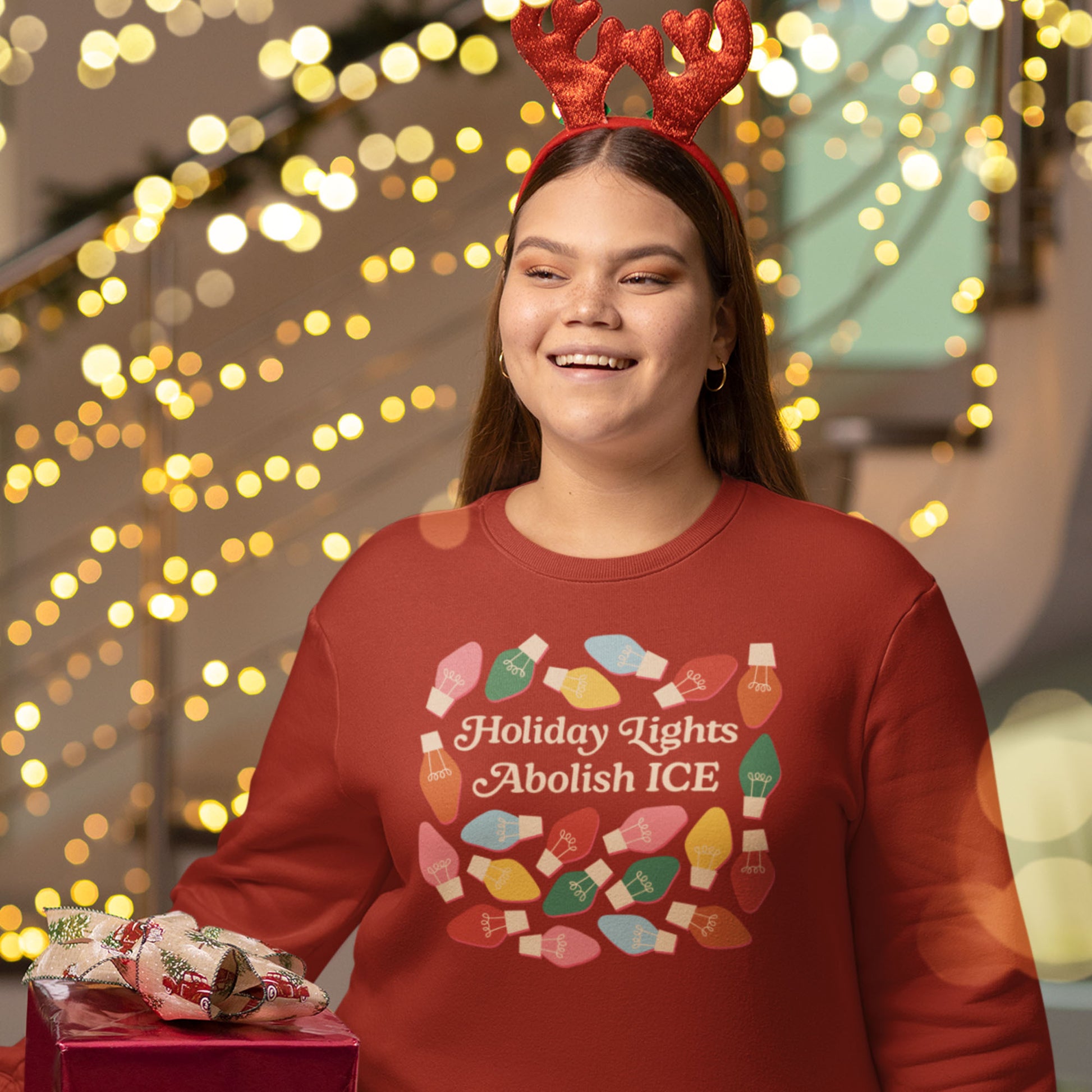 A smiling model wears a red unisex sweatshirt featuring colorful holiday bulbs and “Holiday Lights Abolish ICE,” captured in a cozy Christmas setting with soft glowing lights.