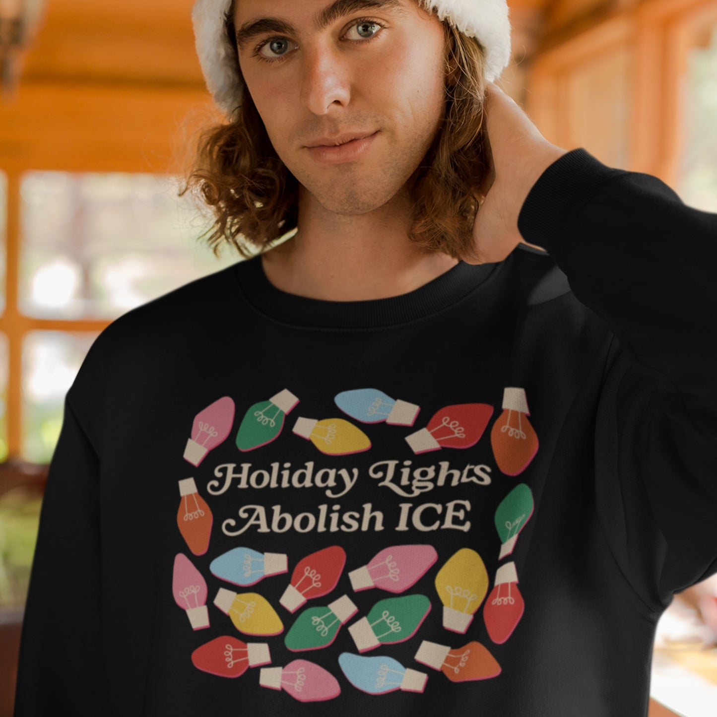 A model wears a black unisex sweatshirt printed with festive holiday bulbs around “Holiday Lights Abolish ICE,” styled in a warm indoor setting for a justice-themed holiday look from Blue Wave USA.