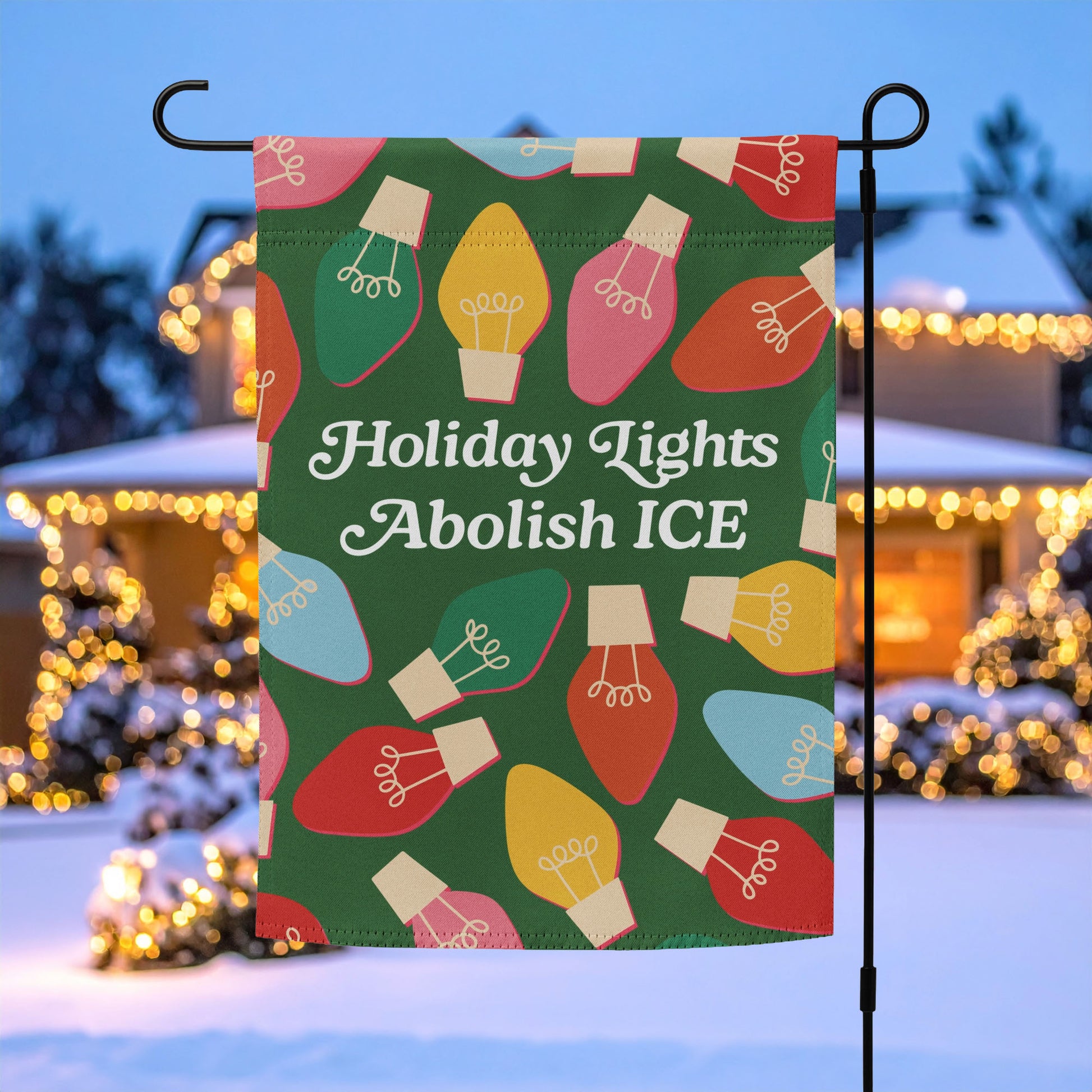 A green garden flag printed with colorful holiday bulbs and the message “Holiday Lights Abolish ICE,” displayed on a flagpole outside a home decorated with warm Christmas lights.
