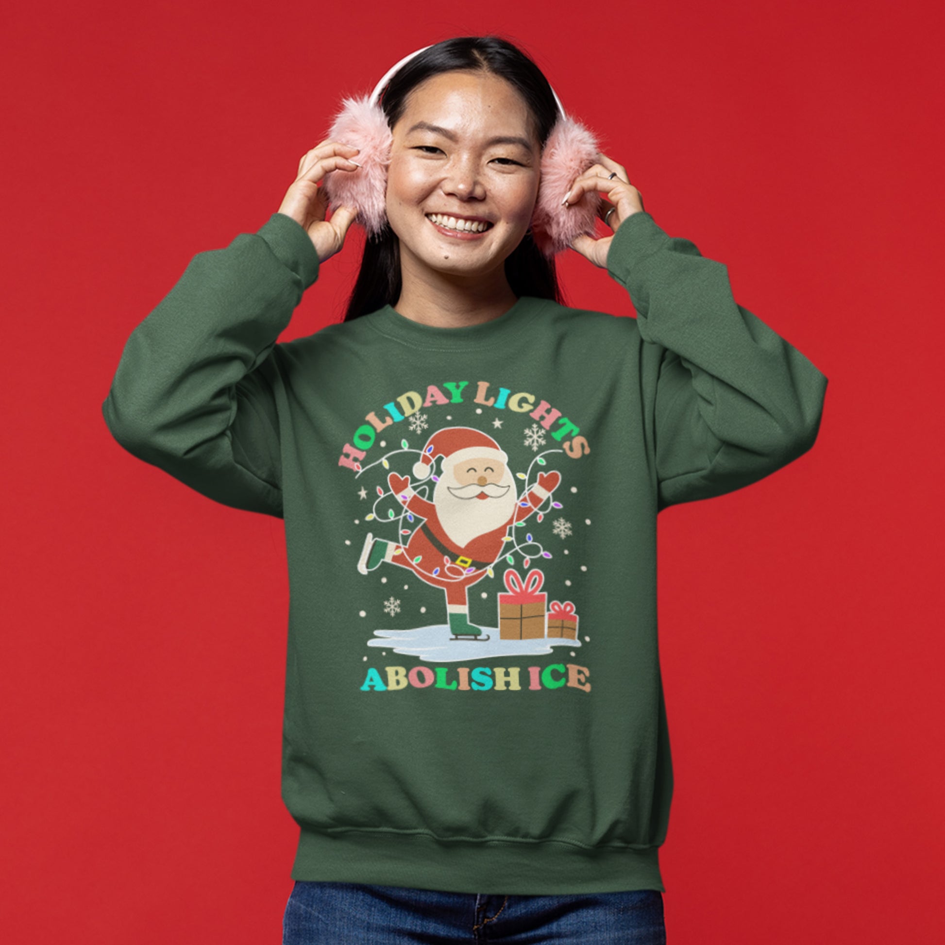 A smiling model wearing a green unisex sweatshirt featuring Santa, festive lights, stars, and presents arranged around the phrase “HOLIDAY LIGHTS ABOLISH ICE,” perfect for playful activist holiday energy.
