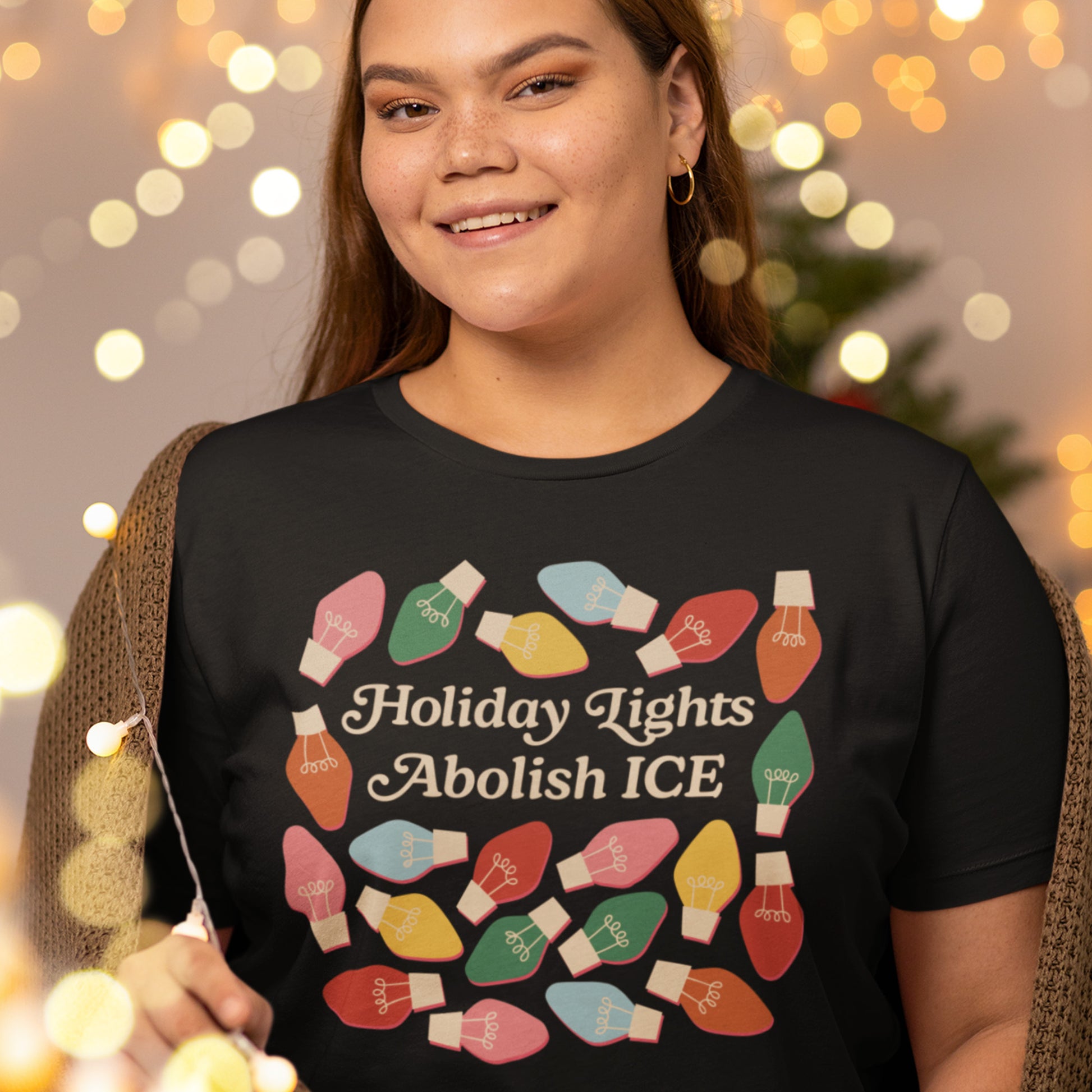 A smiling model wears a black unisex t-shirt decorated with bright holiday bulbs around the message “Holiday Lights Abolish ICE,” shown in warm festive lighting for Blue Wave USA.