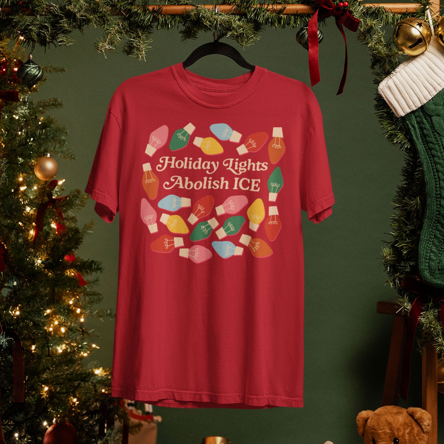 A red unisex t-shirt hangs beside a Christmas tree, featuring colorful holiday bulbs around the message “Holiday Lights Abolish ICE,” styled for progressive holiday décor.