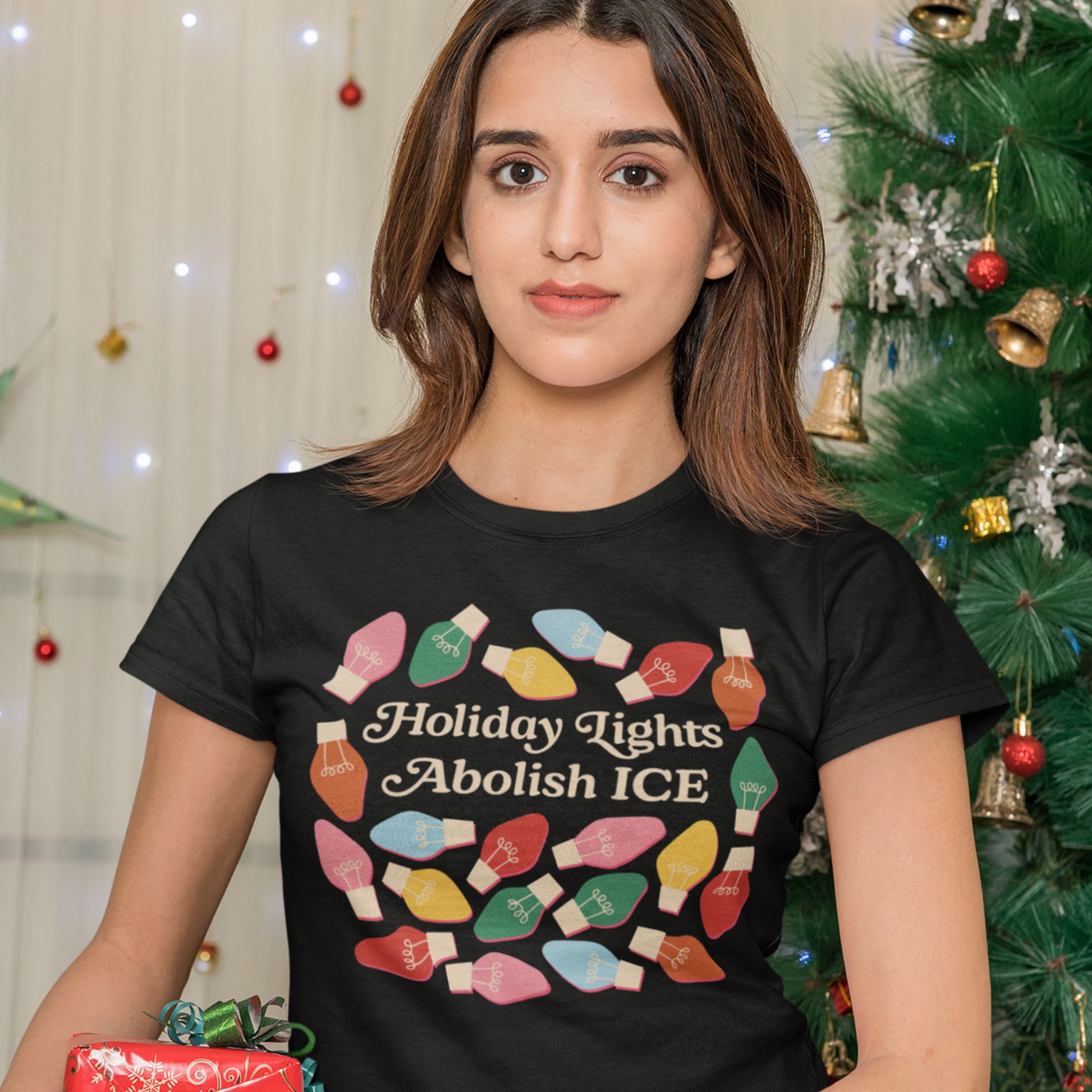 A model wears a black Women’s Relaxed T-Shirt decorated with bright holiday bulbs around “Holiday Lights Abolish ICE,” posed in a soft holiday setting by Blue Wave USA.