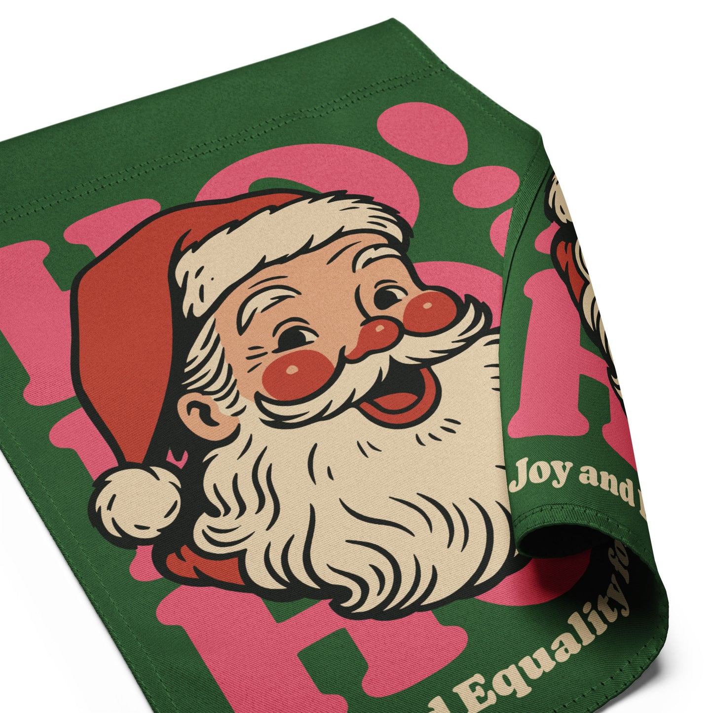 Close-up angled shot of the green “HO! Joy and Equality for All” holiday garden flag, showing a detailed view of the retro Santa illustration with rosy cheeks, a vintage-style beard, and pink lettering. The edge of the fabric is folded to showcase the stitching and durable material.